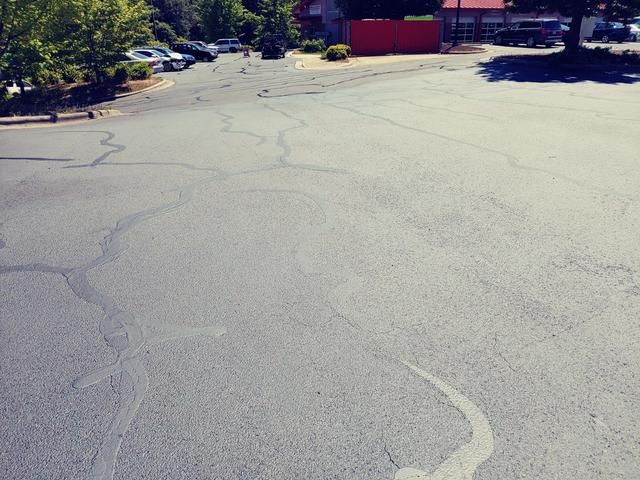 Cracked asphalt parking lot in front of a red building and parked cars under a bright sky.