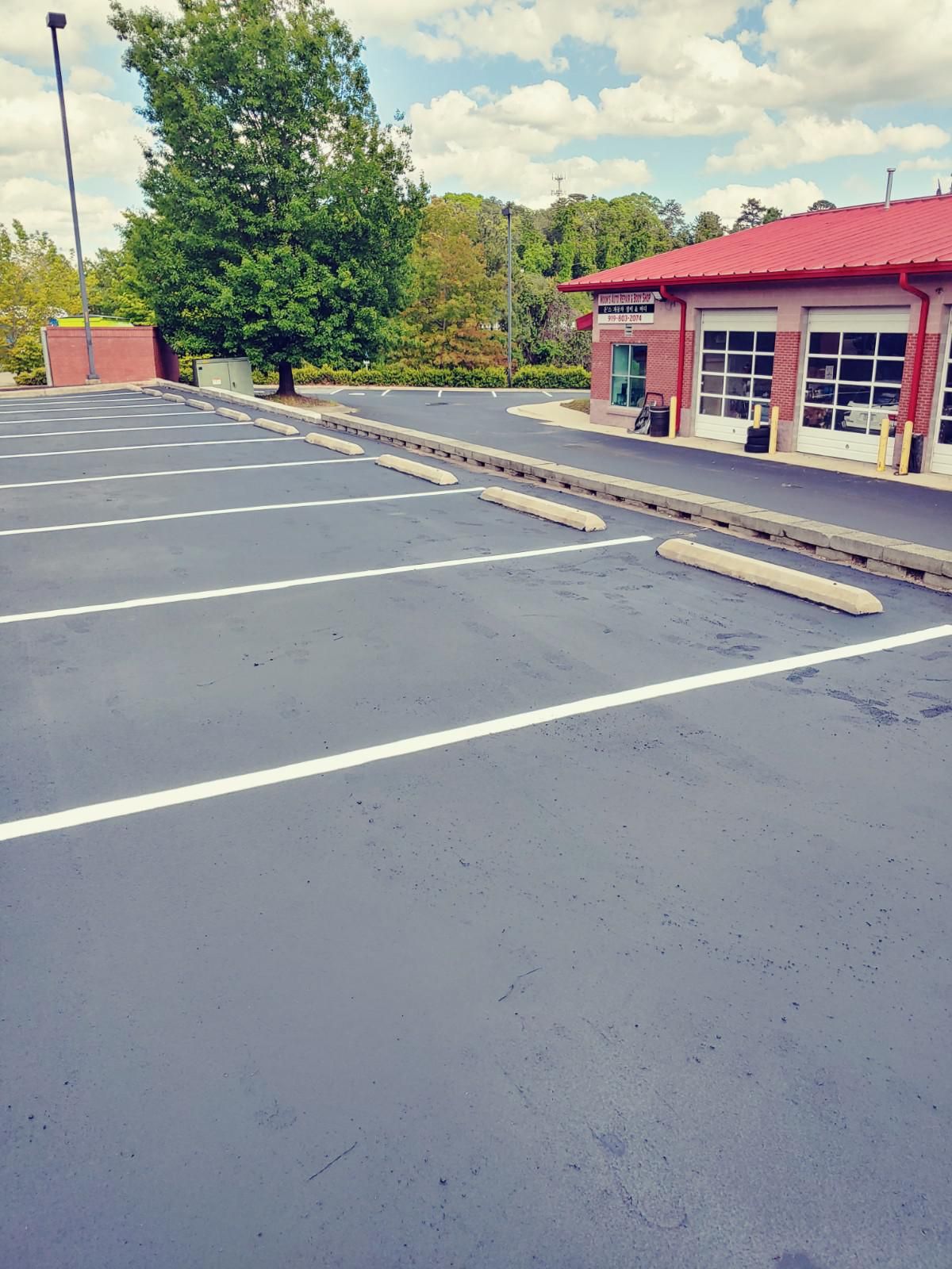 Empty paved parking lot with white lines and wheel stops, building with red roof in background.