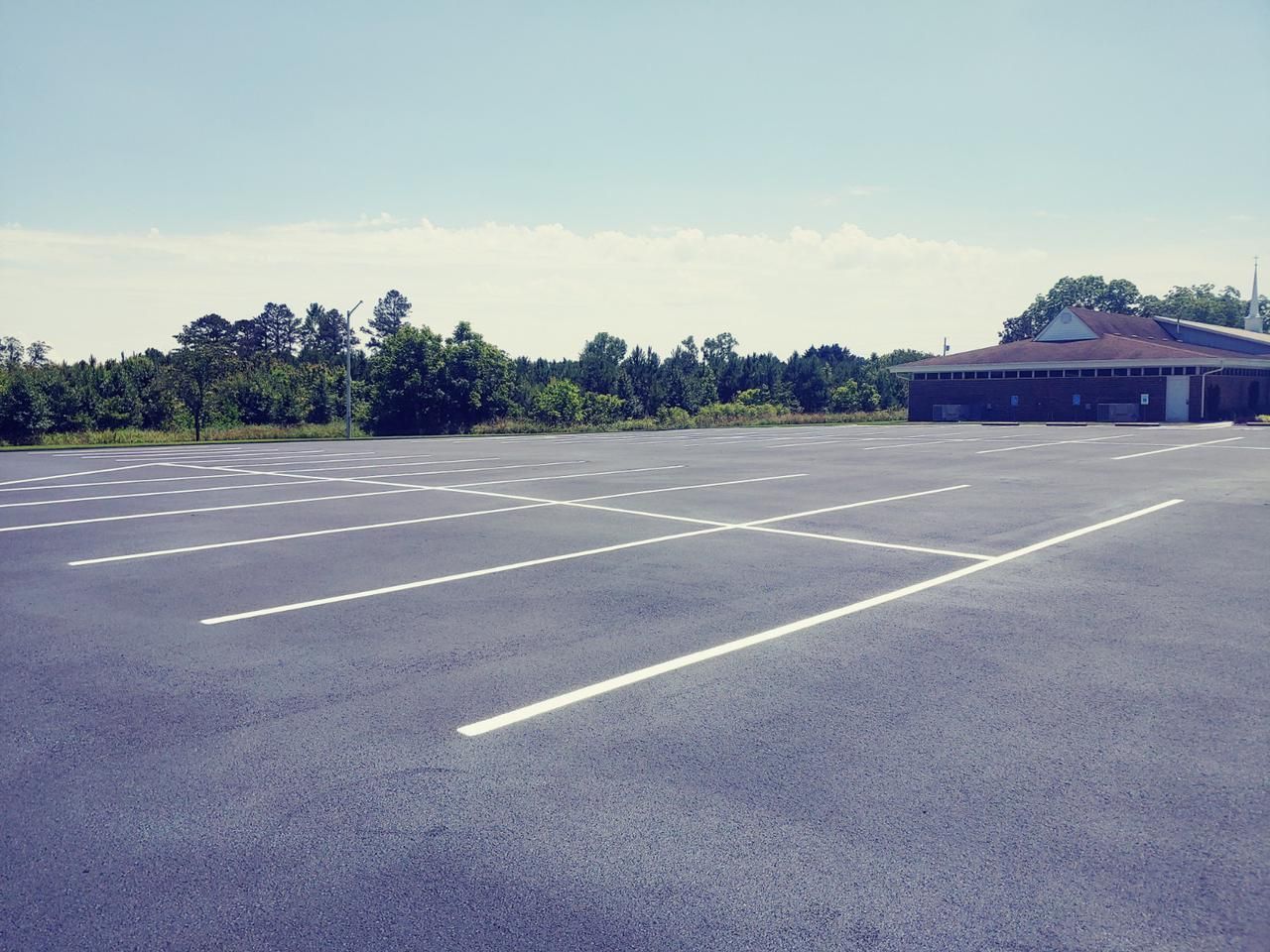 Empty asphalt parking lot with white painted lines, building in background, trees and blue sky.