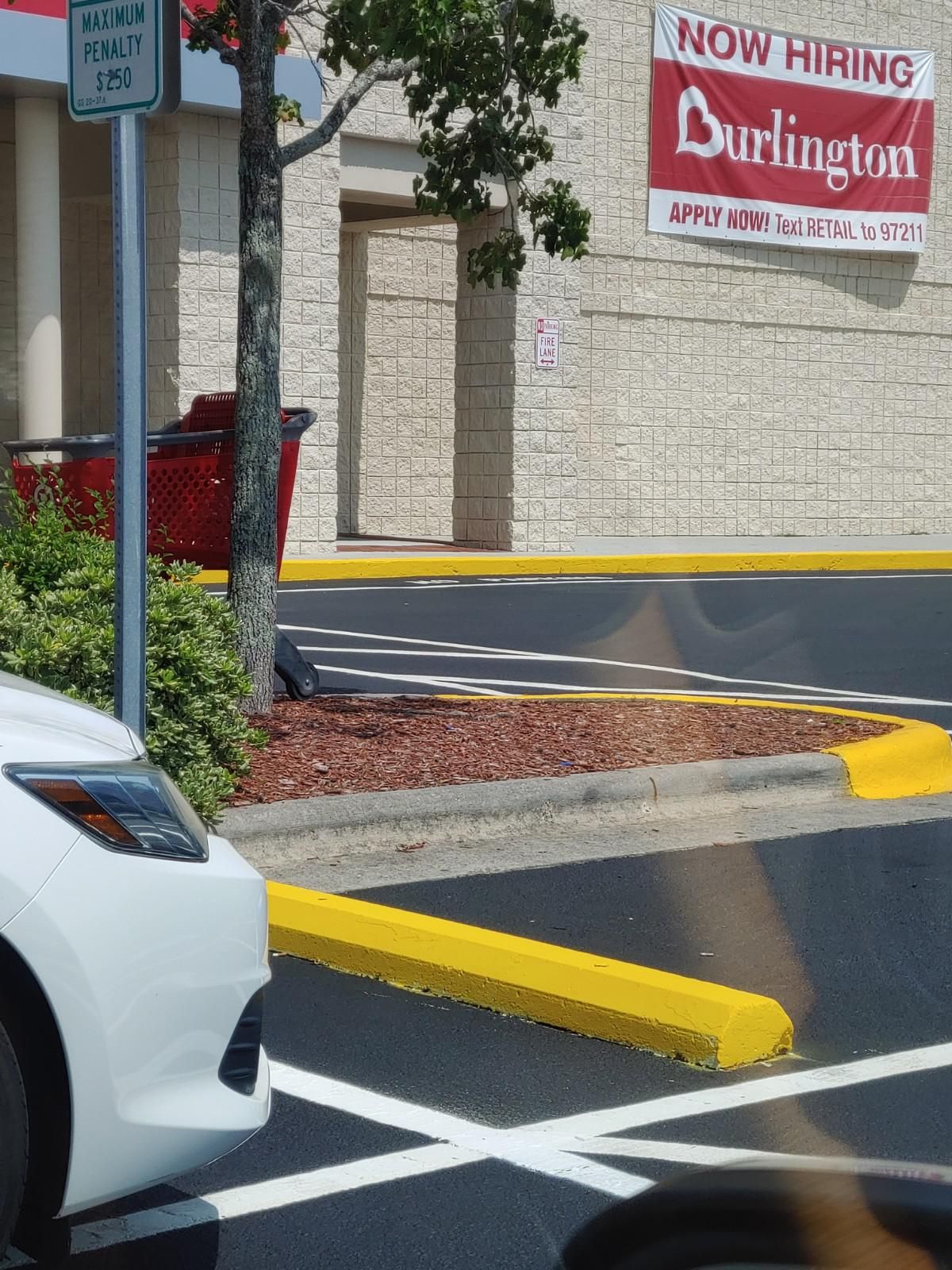White car near a Burlington store entrance. Yellow curb, tree, and “Now Hiring” sign.