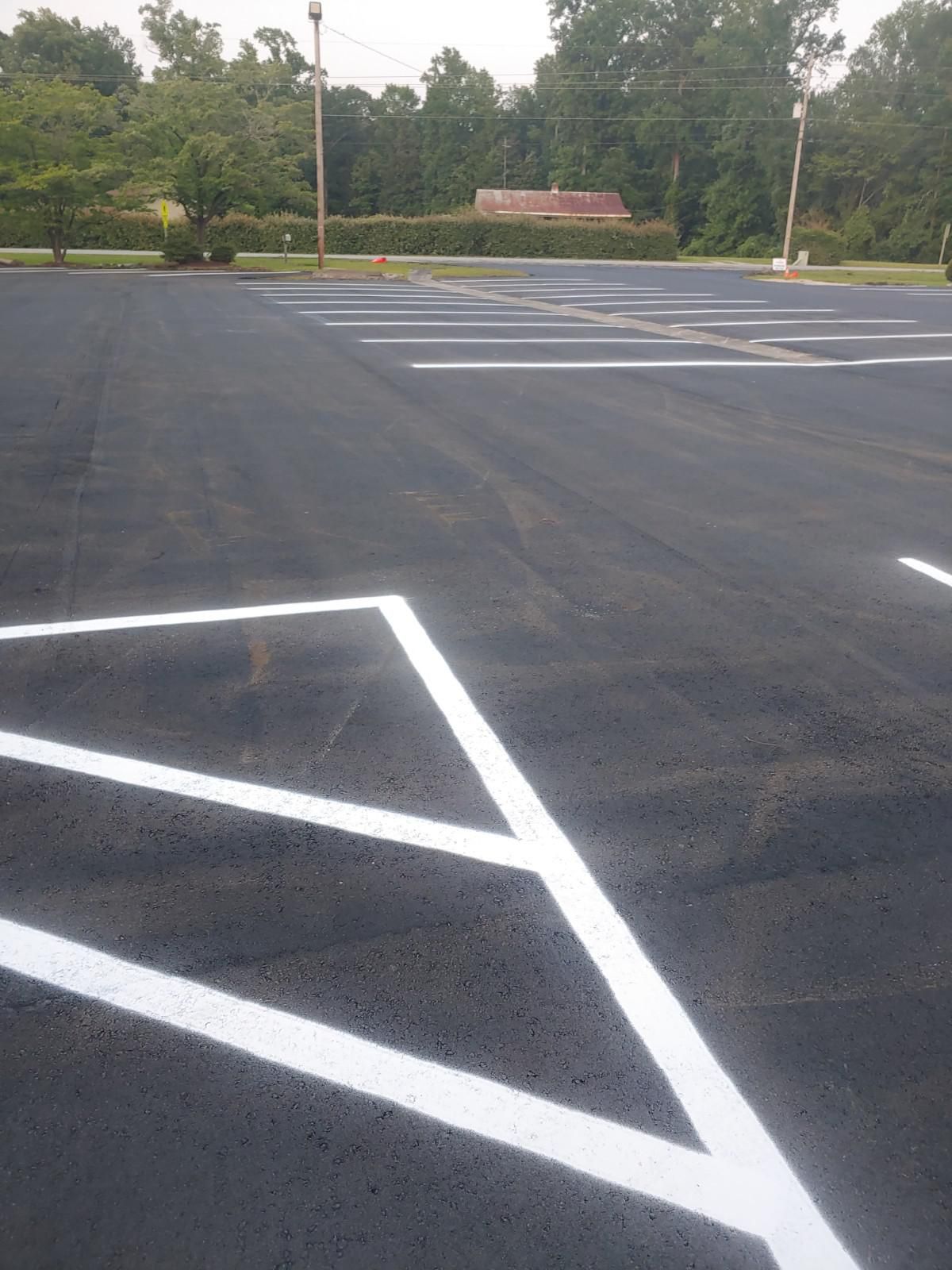 Newly paved parking lot with white painted parking space lines. Trees and a brick building in the background.