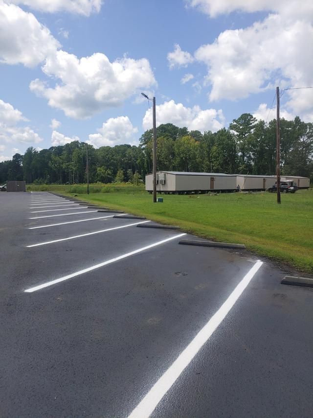 Empty asphalt parking lot with white lines, grass, and trees under a cloudy sky.