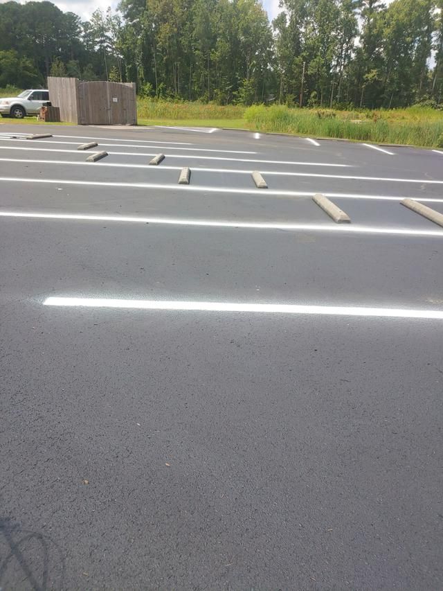 Empty asphalt parking lot with white lines and wheel stops, with a wooden structure and trees in the background.
