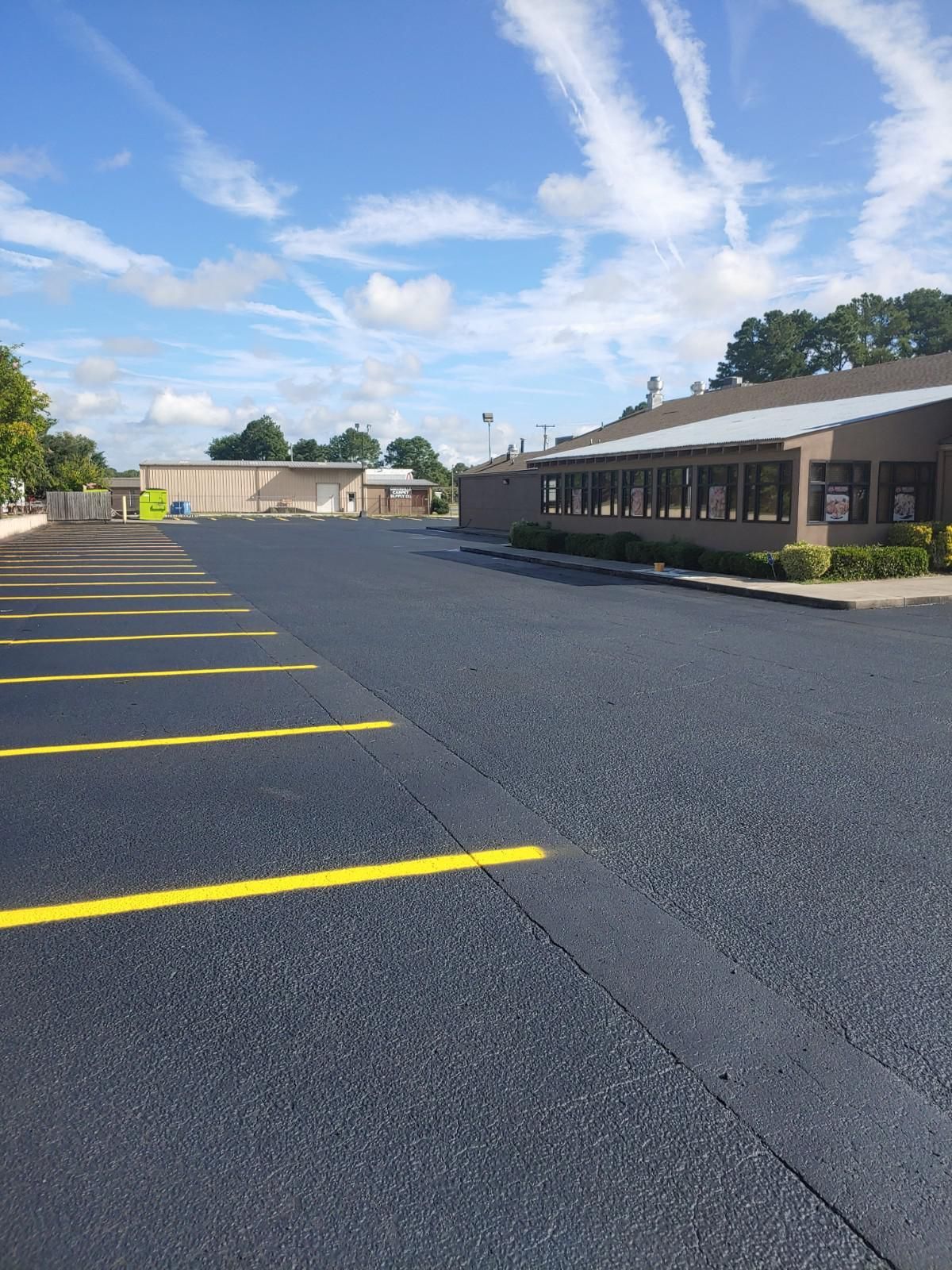 Empty asphalt parking lot with yellow lines, two low buildings, and cloudy sky.