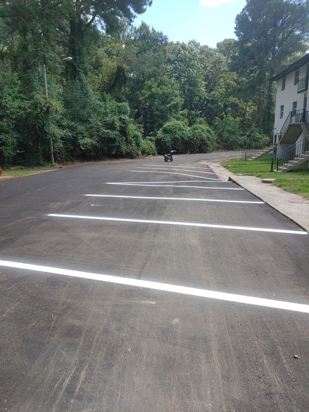Asphalt parking lot with white painted parking space lines, building and trees in background.