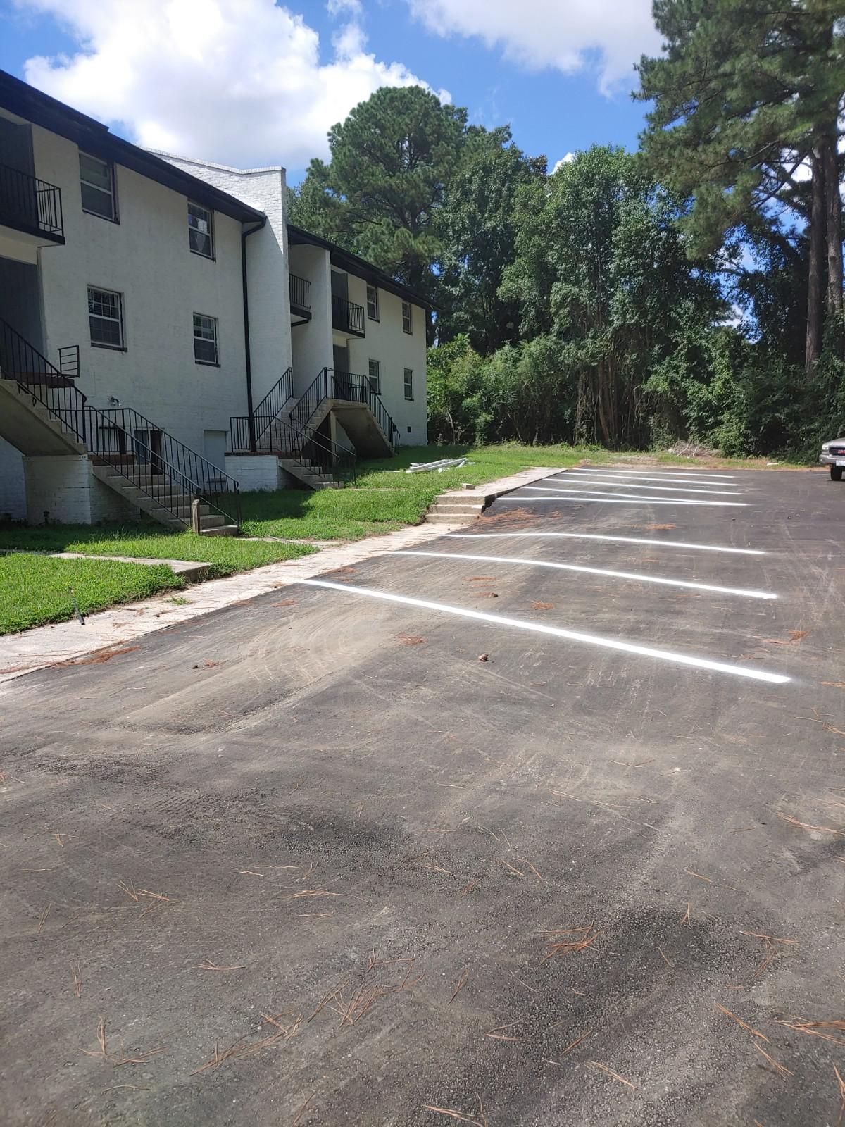Apartment building next to a parking lot under a blue sky, with trees in the background.