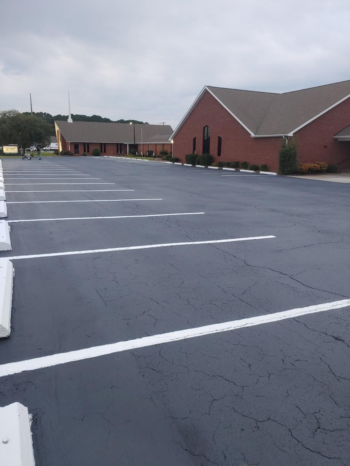 Empty asphalt parking lot with white lines in front of brick church buildings under cloudy sky.
