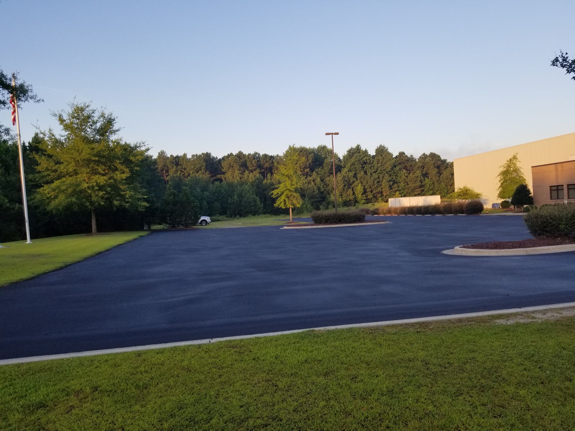 An empty asphalt parking lot surrounded by grass and trees on a sunny day.