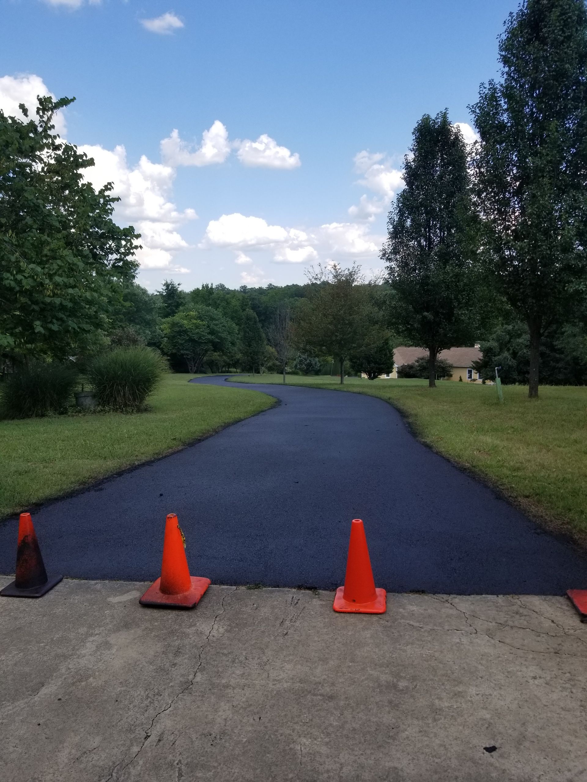 Freshly paved black driveway with orange safety cones, curving toward trees under a blue sky.