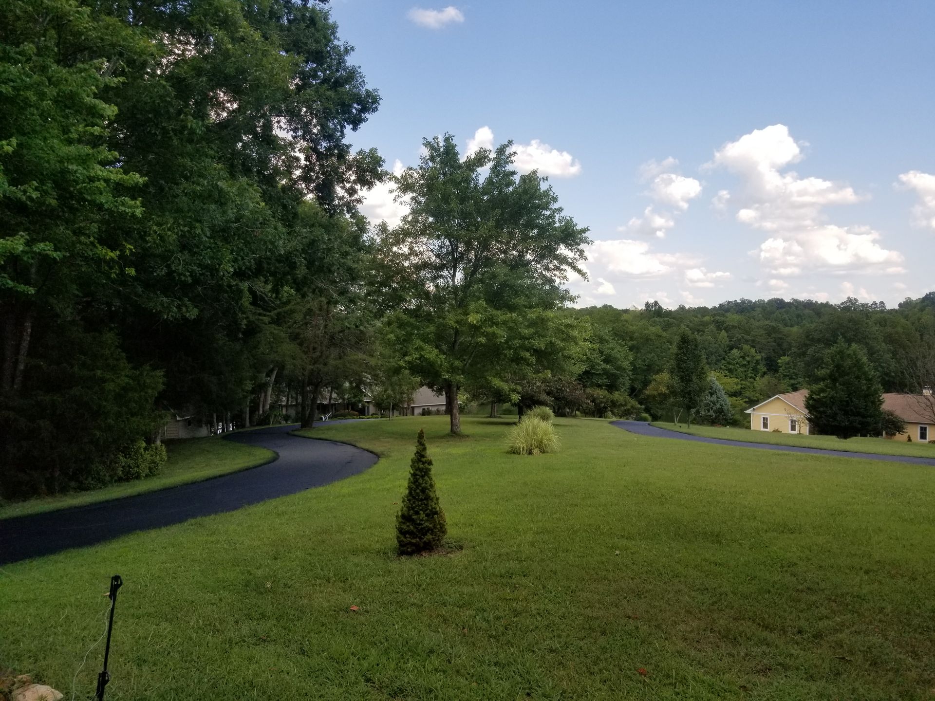 Lush green lawn with a black paved driveway curves towards a distant house under a blue sky with fluffy clouds.