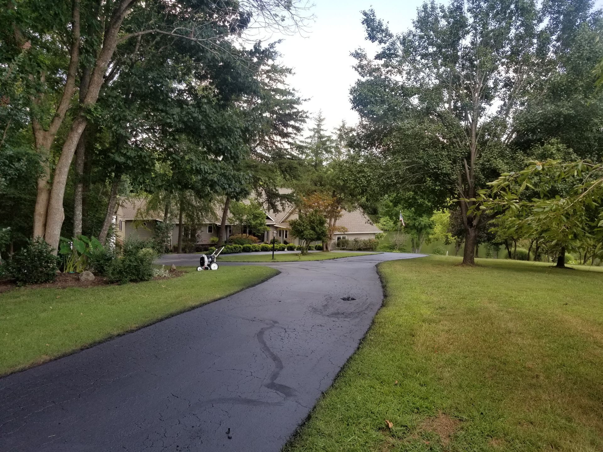 A long, black driveway winds through a green, grassy area toward a house framed by trees.