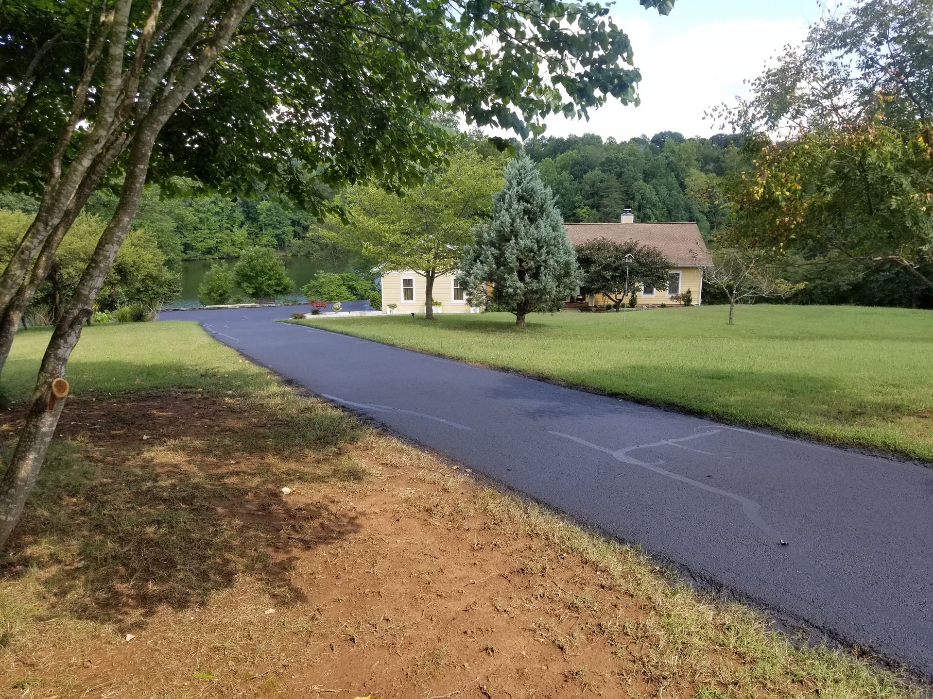 Paved driveway leading to a light-colored house surrounded by green grass and trees on a sunny day.