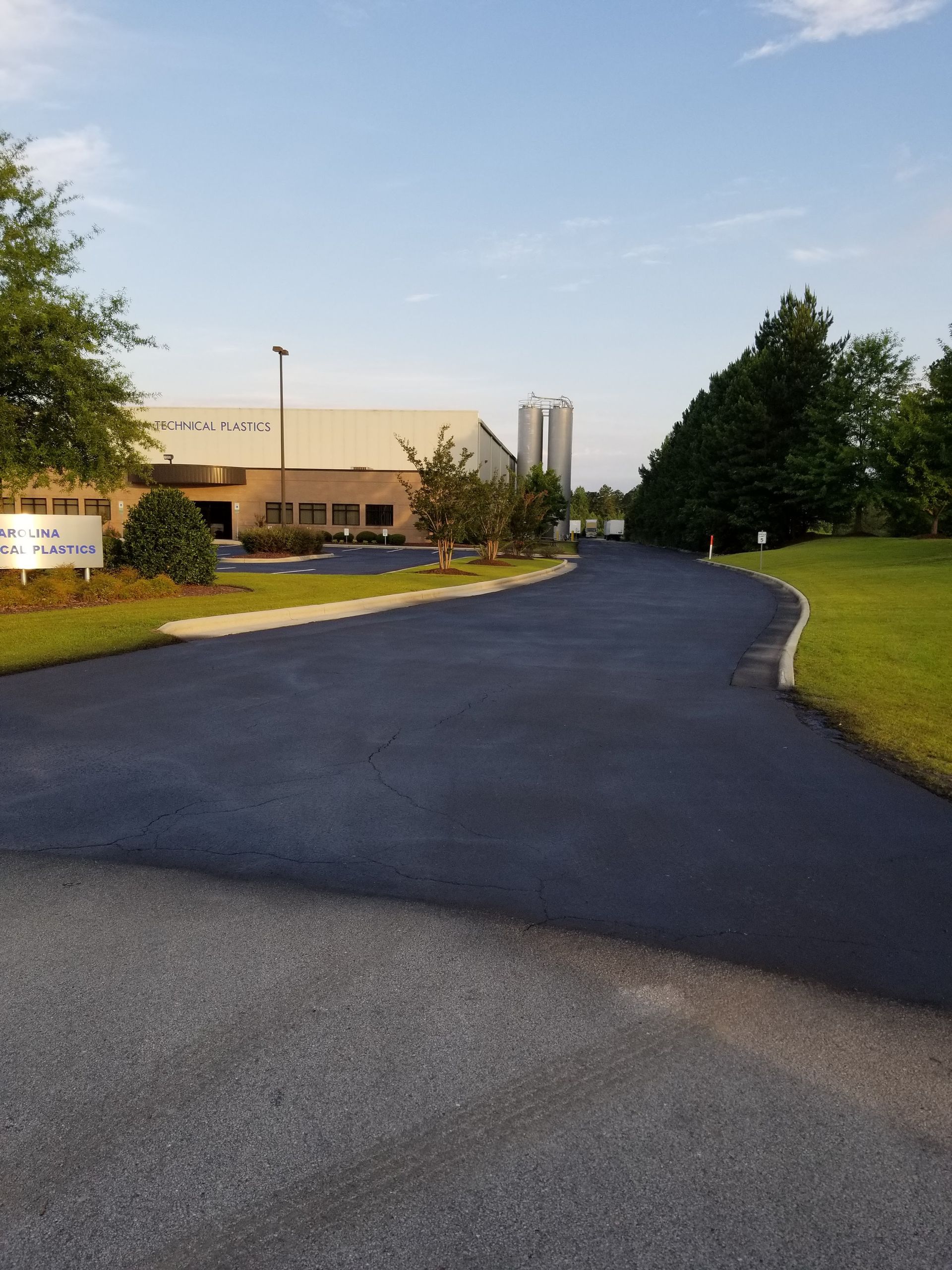 Paved driveway leading toward a low, tan building under a blue sky, with some trees and a tall white silo visible.