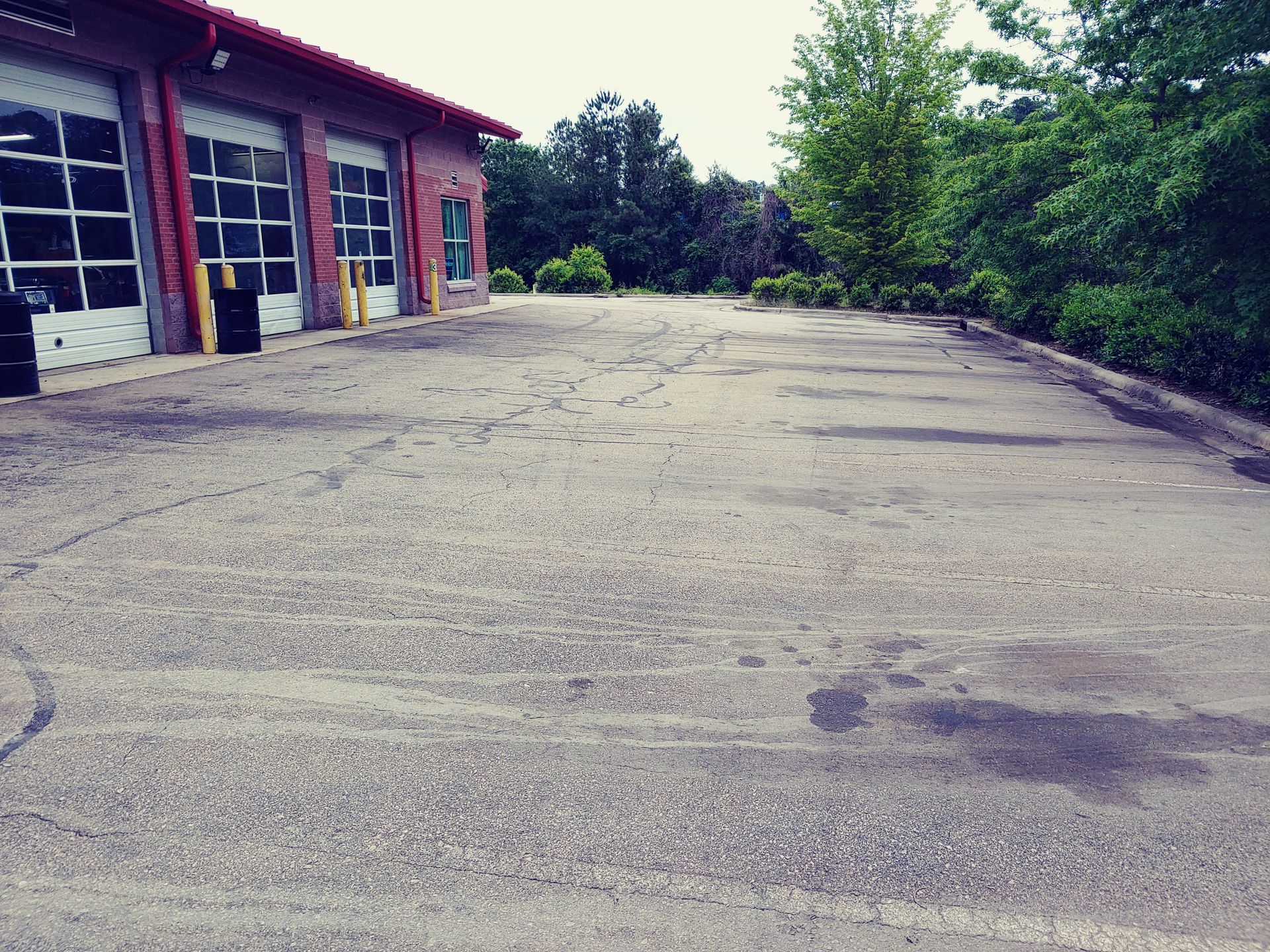 Exterior of a brick building with garage doors and a cracked asphalt parking area, trees in background.