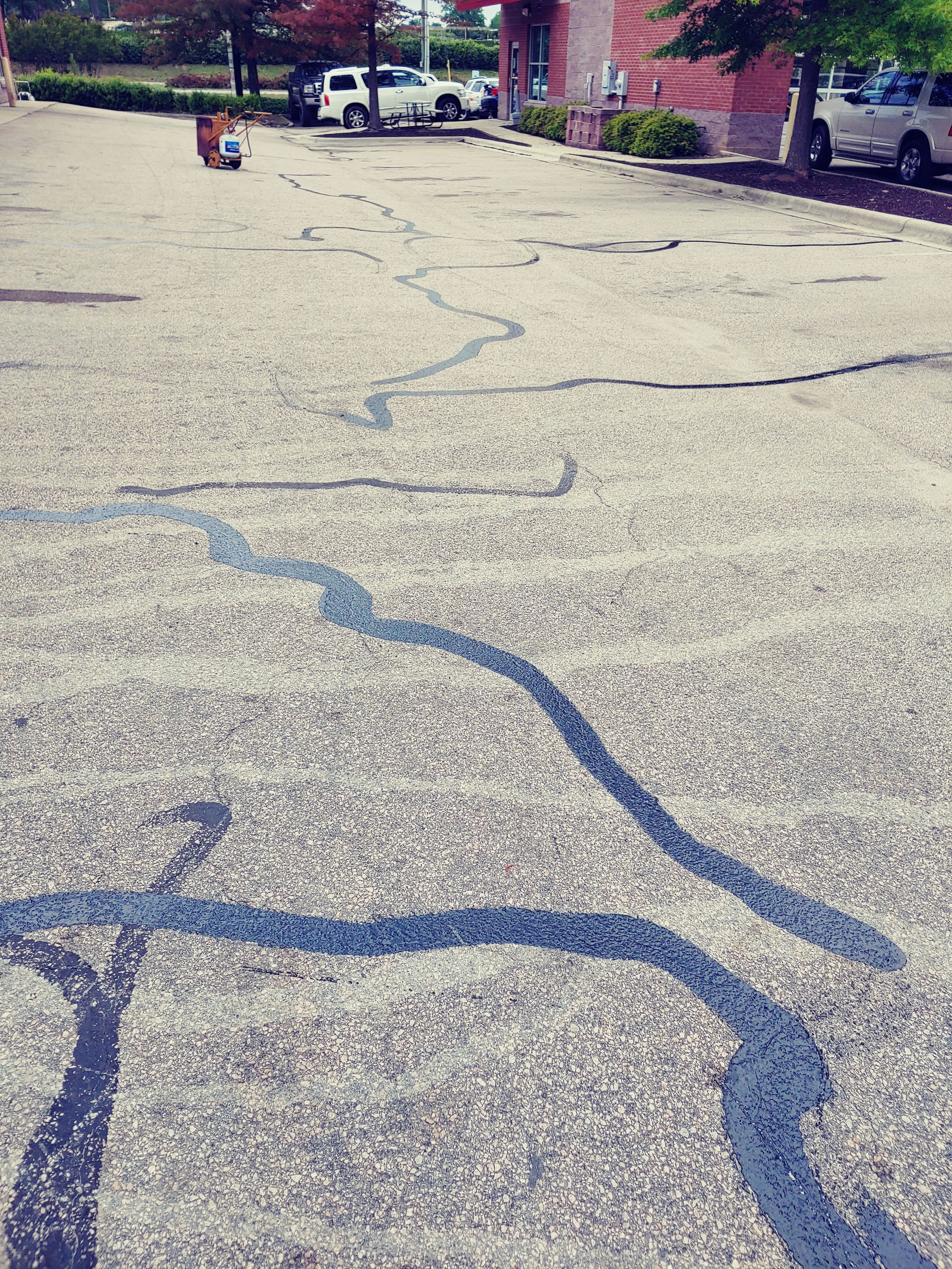 Cracked asphalt parking lot with black sealant, vehicles, and a building in the background.