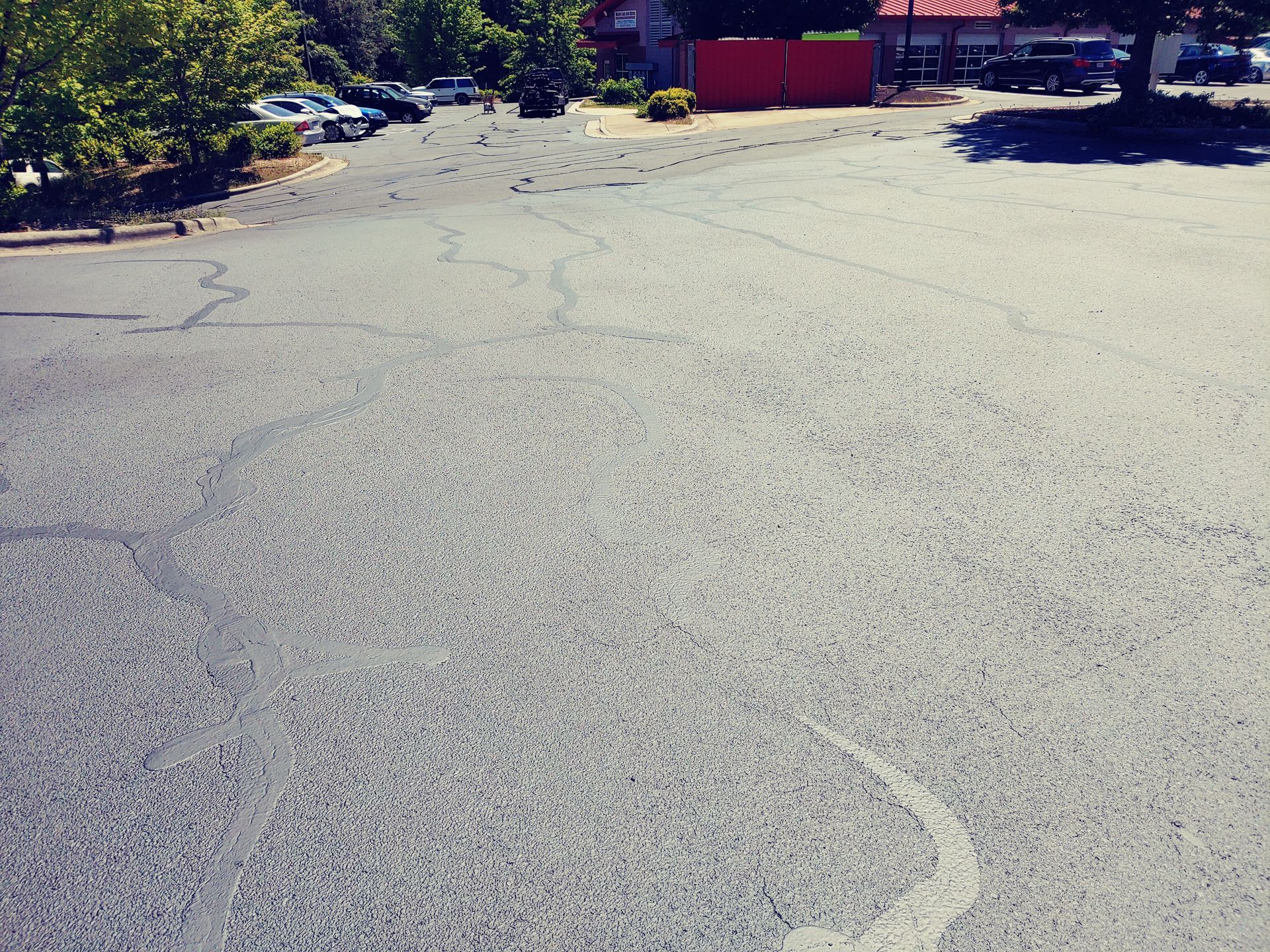 Empty asphalt parking lot near a building with some parked cars. Trees and shadows visible.