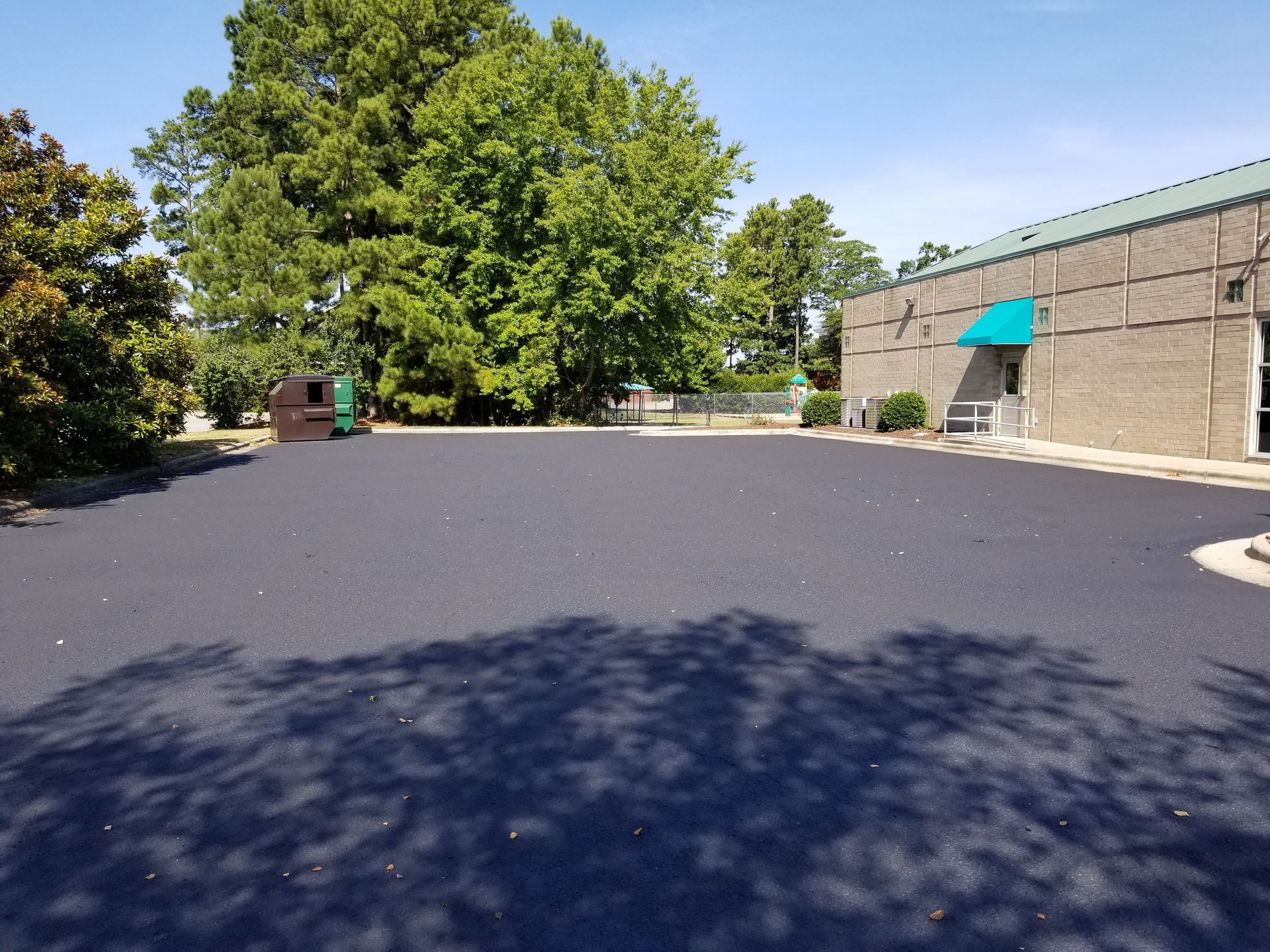 Newly paved asphalt parking lot next to a building with a green awning and trees under a clear sky.