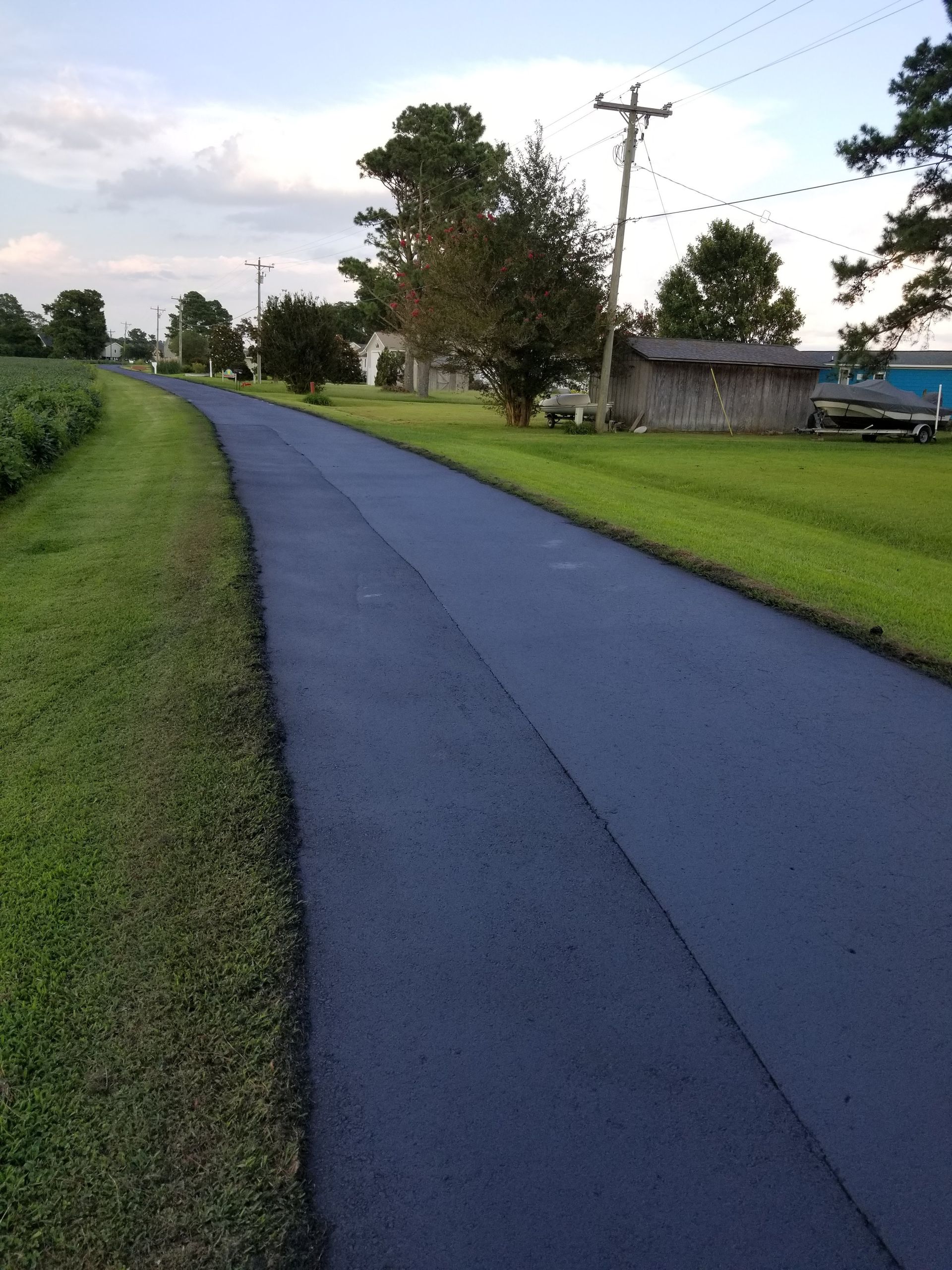 Dark asphalt road curves through a rural area, flanked by green grass and a field. Power lines and trees in the background.