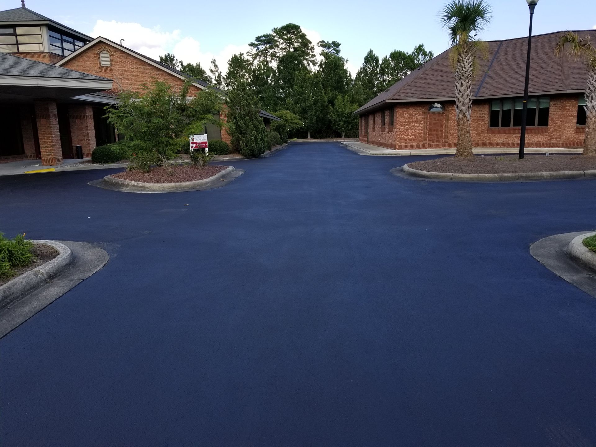 Newly paved dark asphalt driveway in front of brick buildings, trees in the background.