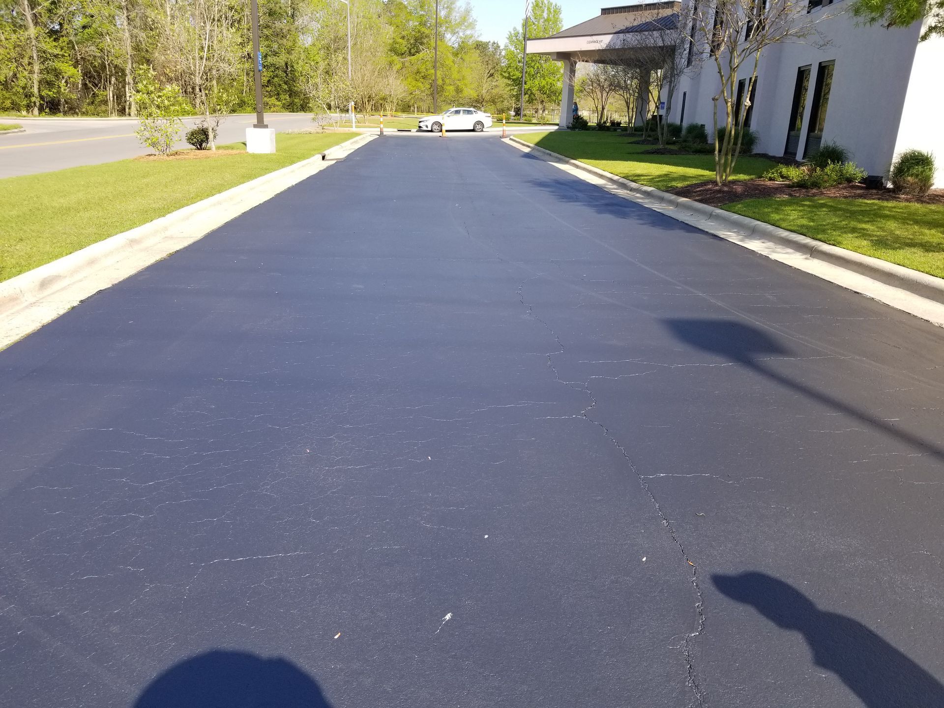 Black asphalt driveway leading to a building, sunlight, white car in the distance.