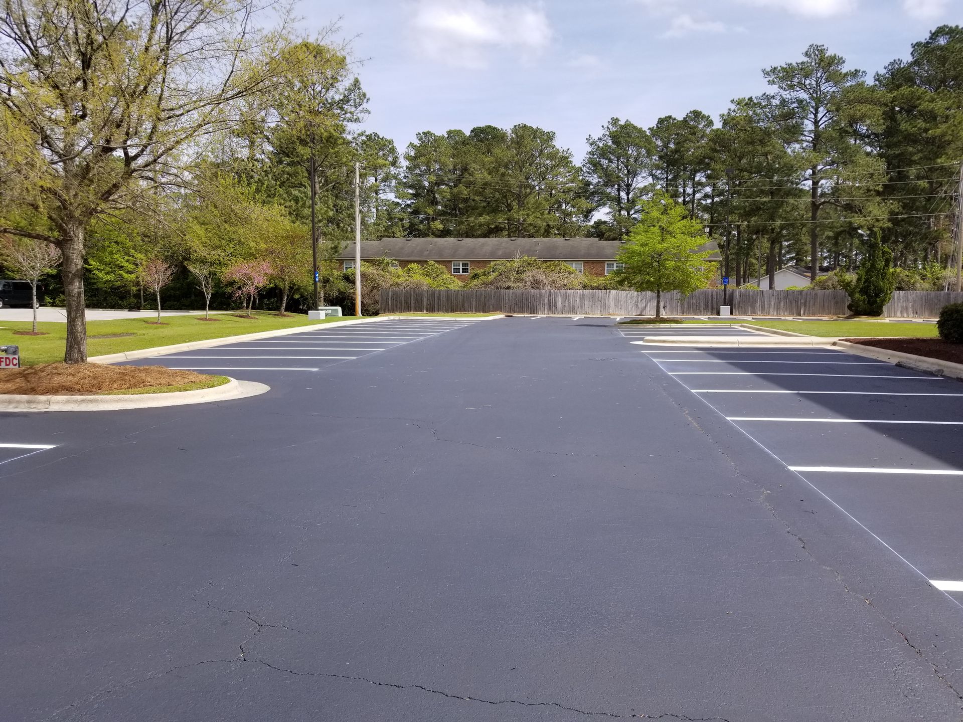 Empty asphalt parking lot with white painted lines; trees and fence in background.