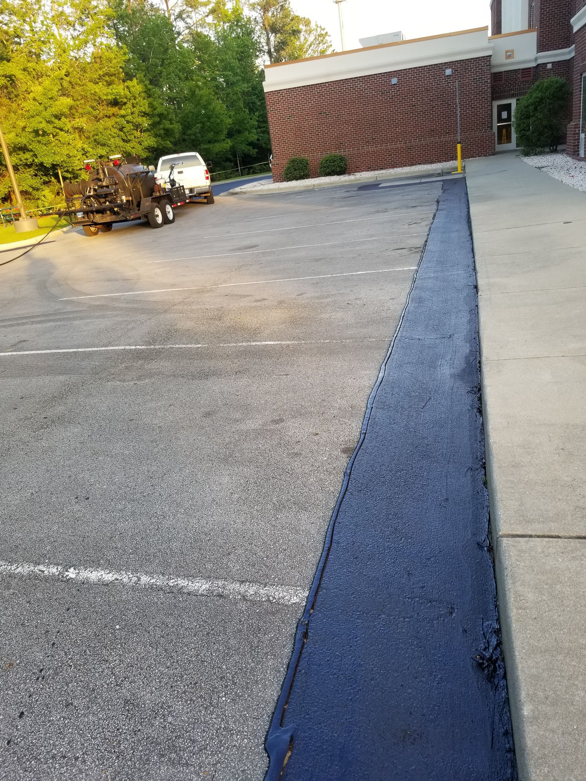 Dark asphalt seal coat application along a concrete edge in a parking lot.