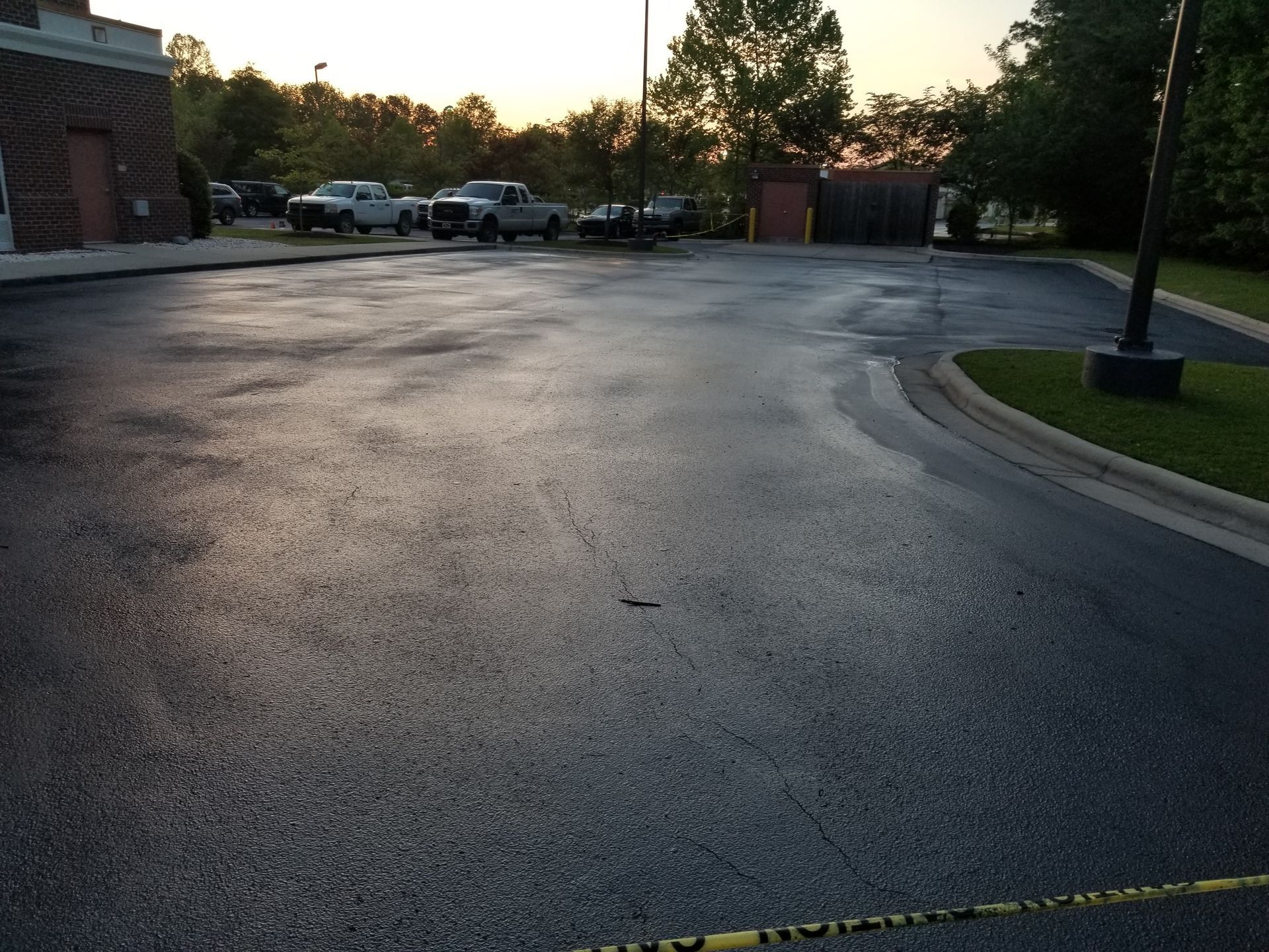 Asphalt parking lot, freshly sealed, reflecting the sky. Vehicles and a building are in the background.