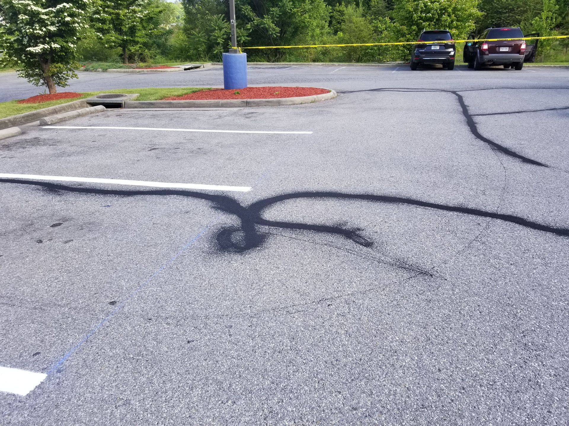 Black skid marks on asphalt parking lot. Two cars, crime scene tape, and a blue trash can visible in background.