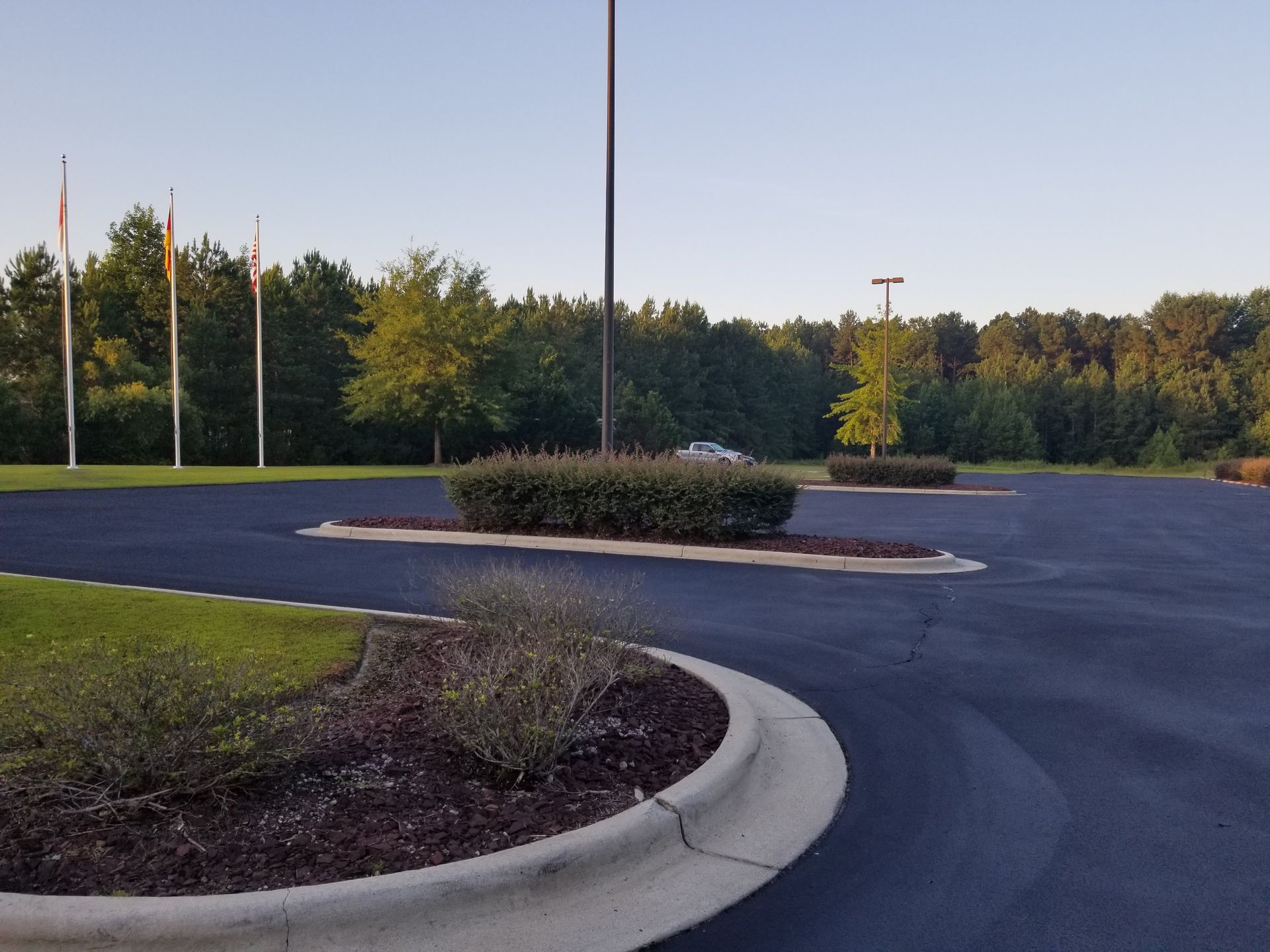 An empty asphalt parking lot with landscaped islands, flags, and trees under a clear sky.