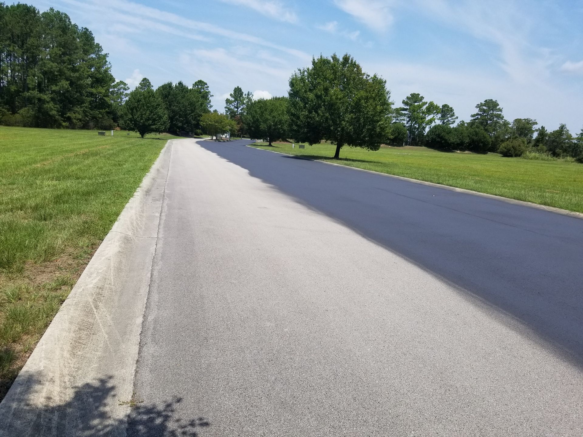 Asphalt road, newly sealed on the right, concrete curb, grassy areas on each side, trees and blue sky.