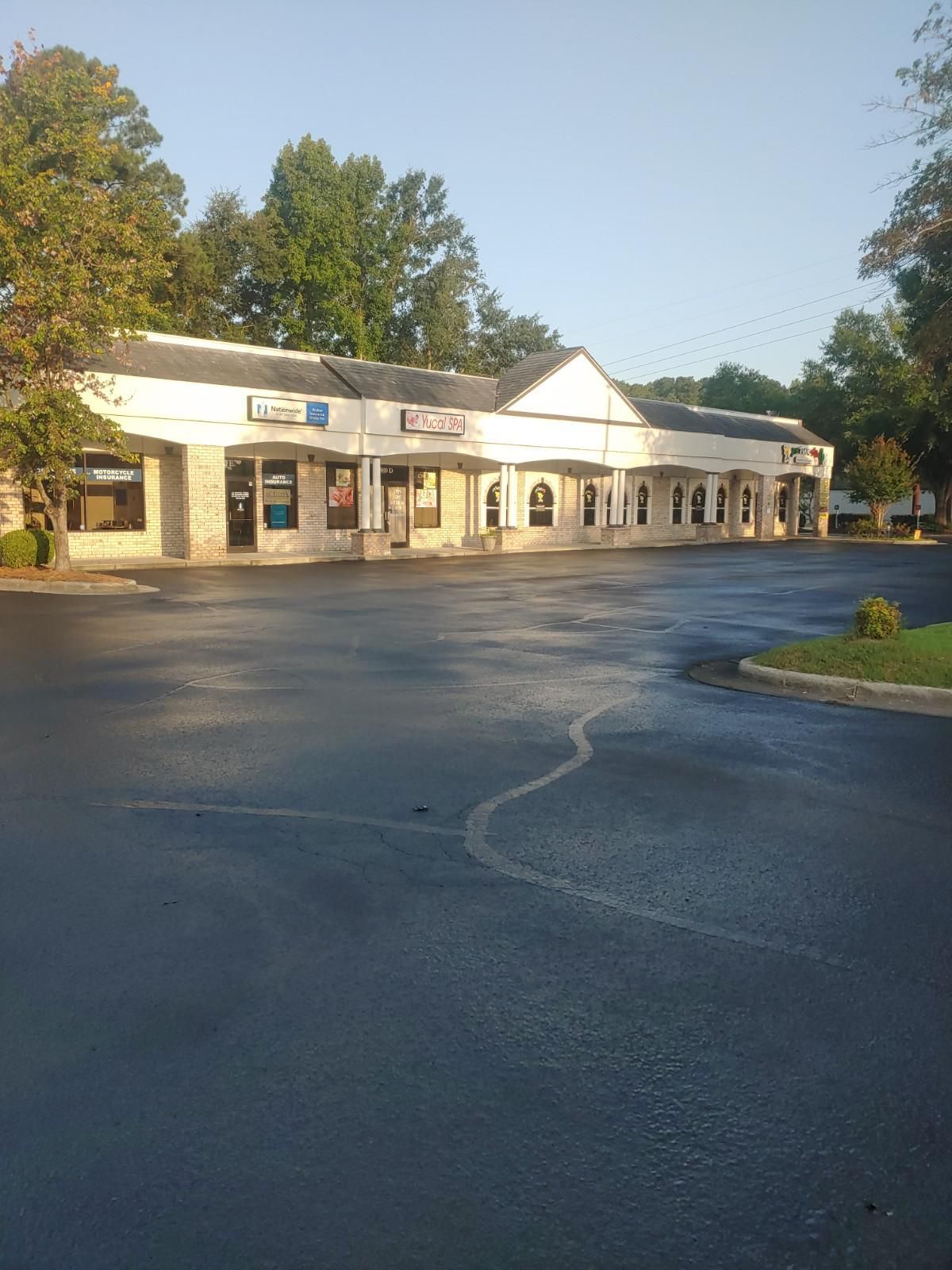 Strip mall with shops along a black asphalt parking lot. Trees in the background.