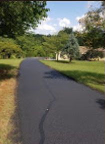 Paved road curves through a green, grassy area, leading toward trees and a clear, blue sky.
