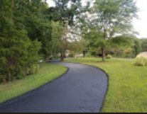 Black asphalt driveway curves through a grassy lawn, lined by trees on either side.