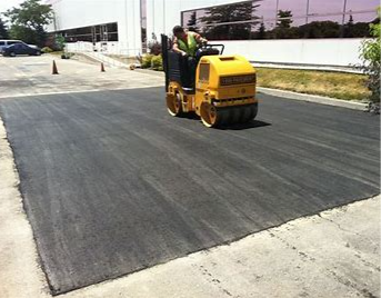 A worker operating a yellow road roller compacting fresh asphalt on a paved surface.