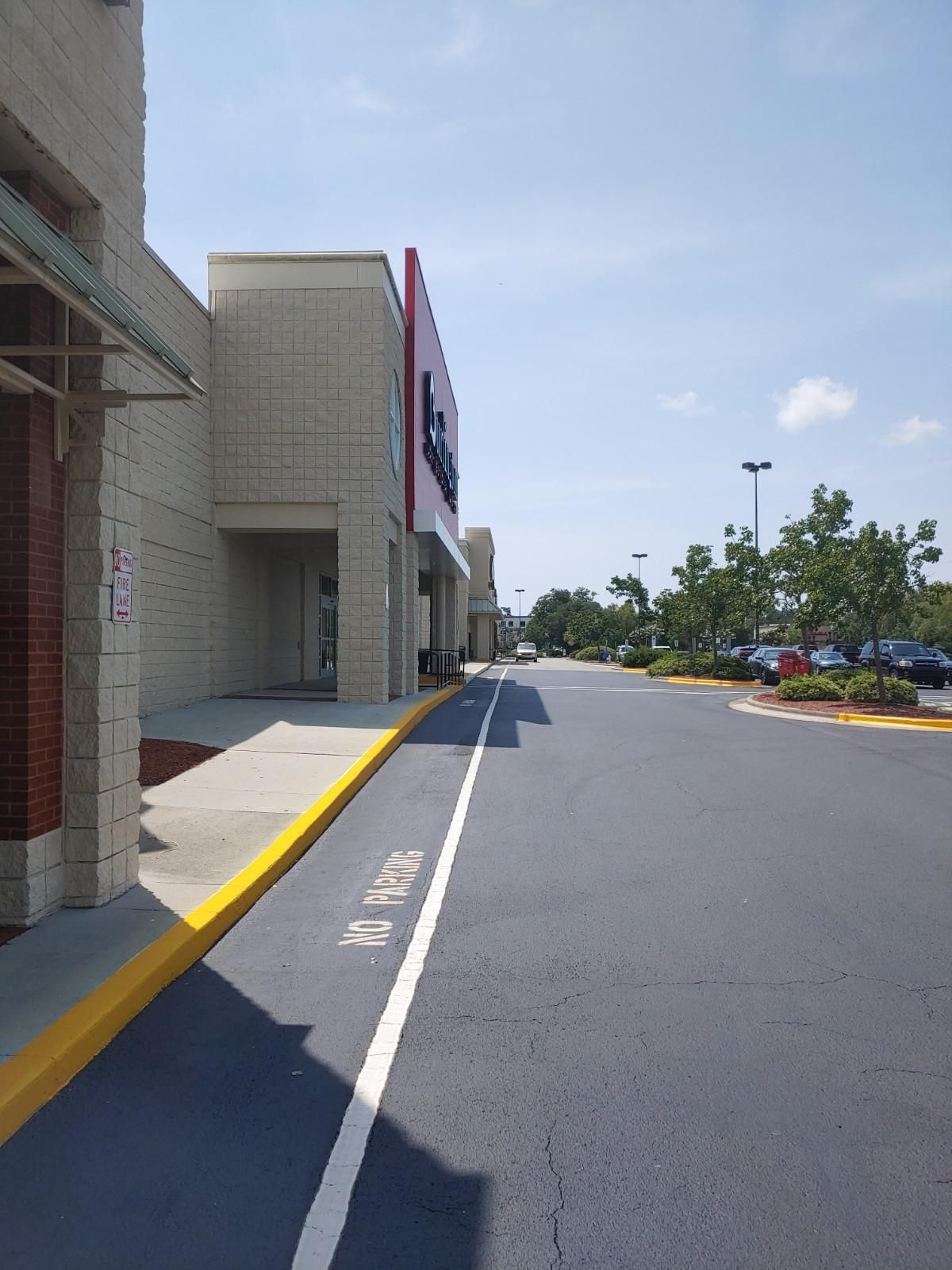 Exterior of a retail store with a paved parking area and blue sky. The building is beige with red signage.