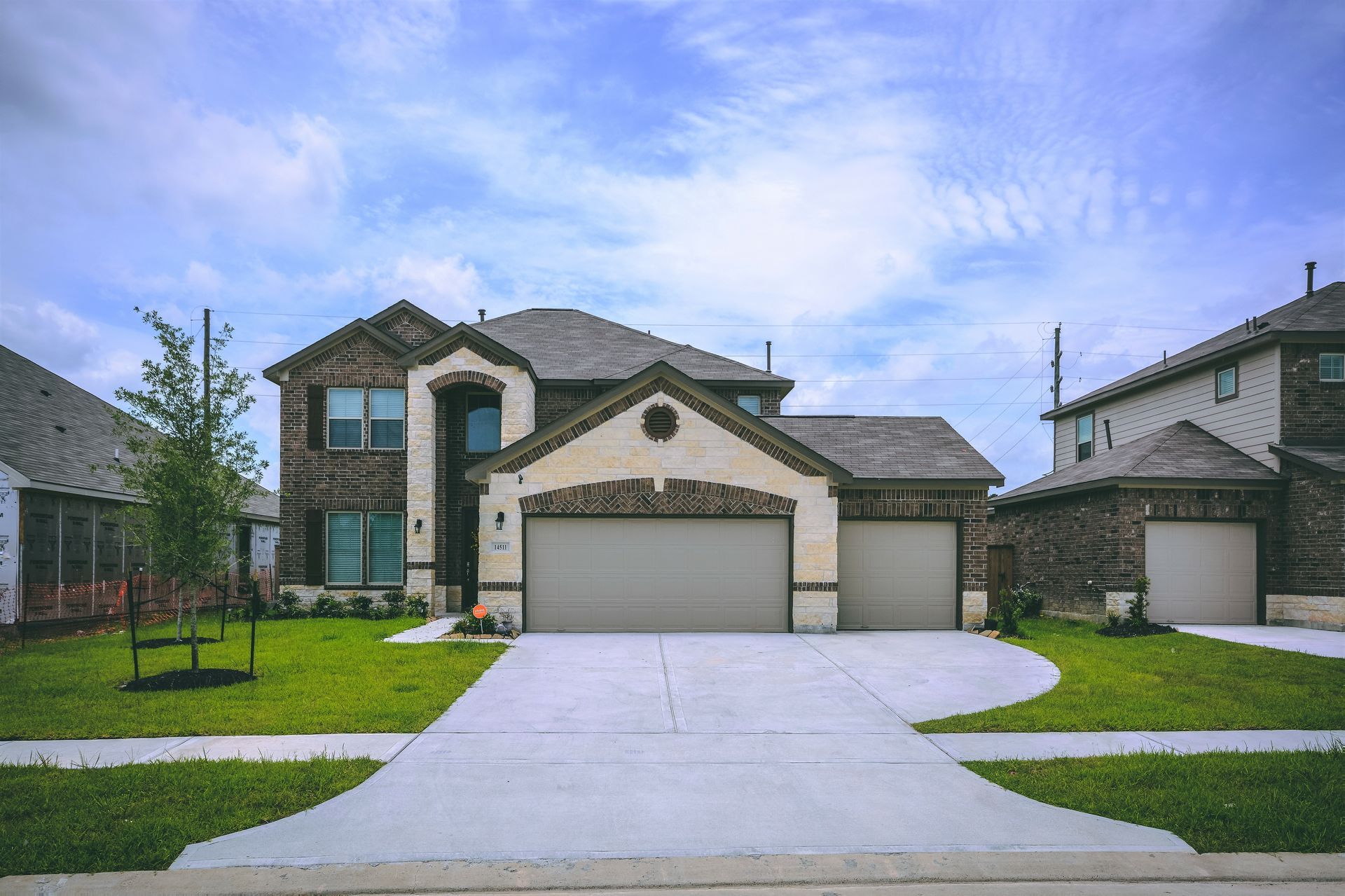 Two-story brick house with attached garage, concrete driveway, and grassy lawn under a blue sky.