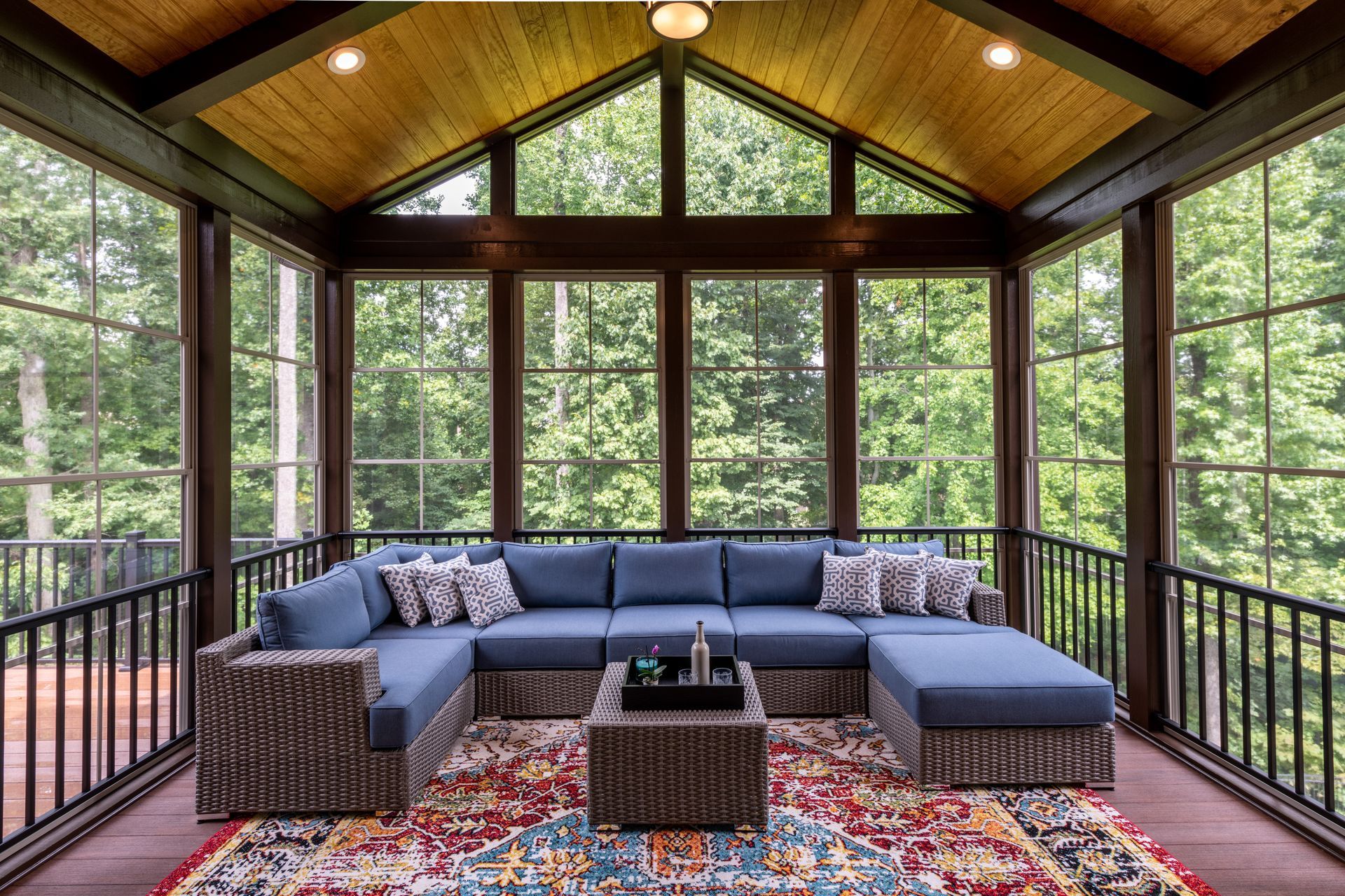 Sunroom with blue sectional sofa, colorful rug, large windows, and wooden ceiling.