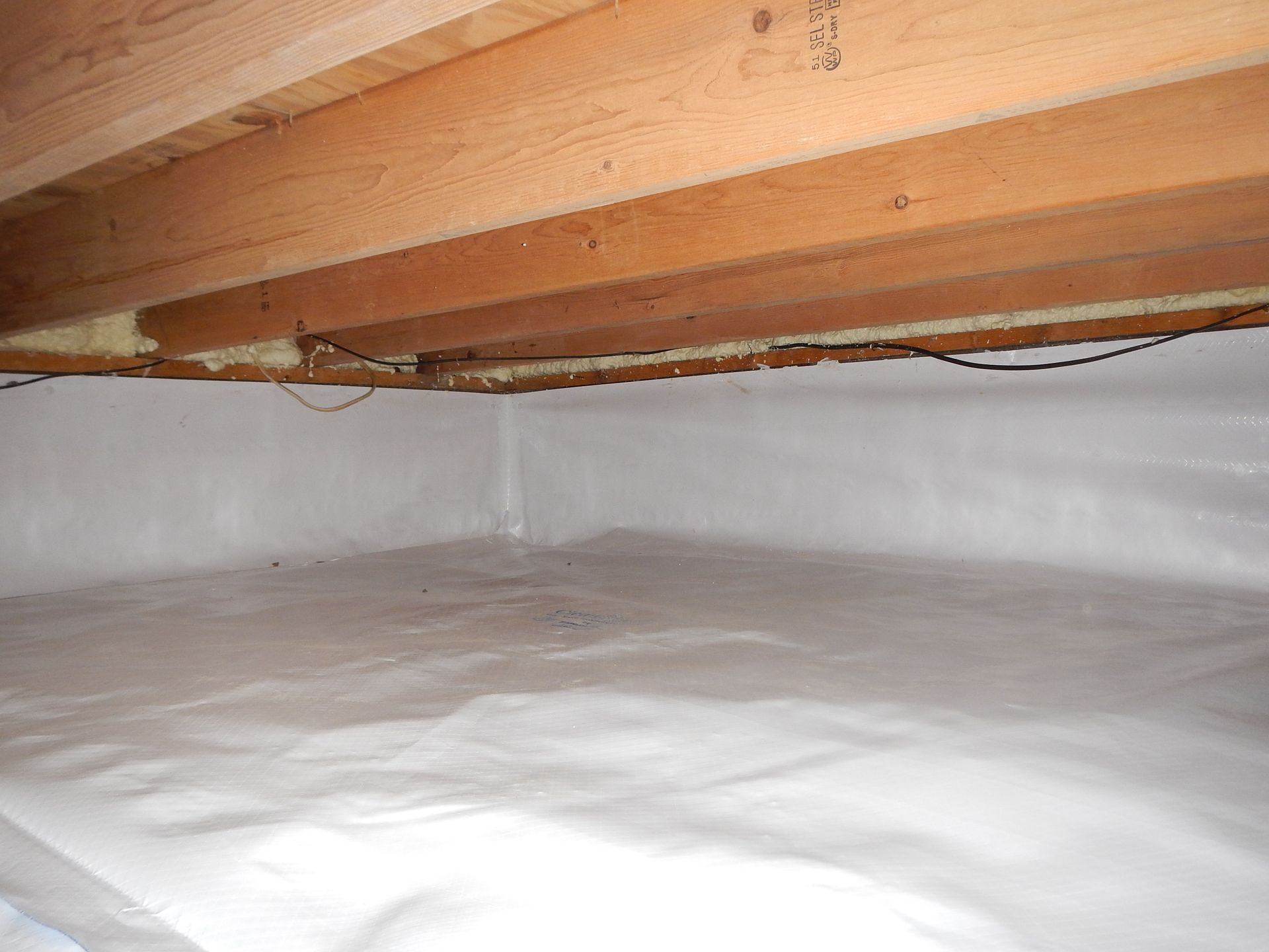 Crawl space with white vapor barrier covering the walls and floor, wooden beams overhead.