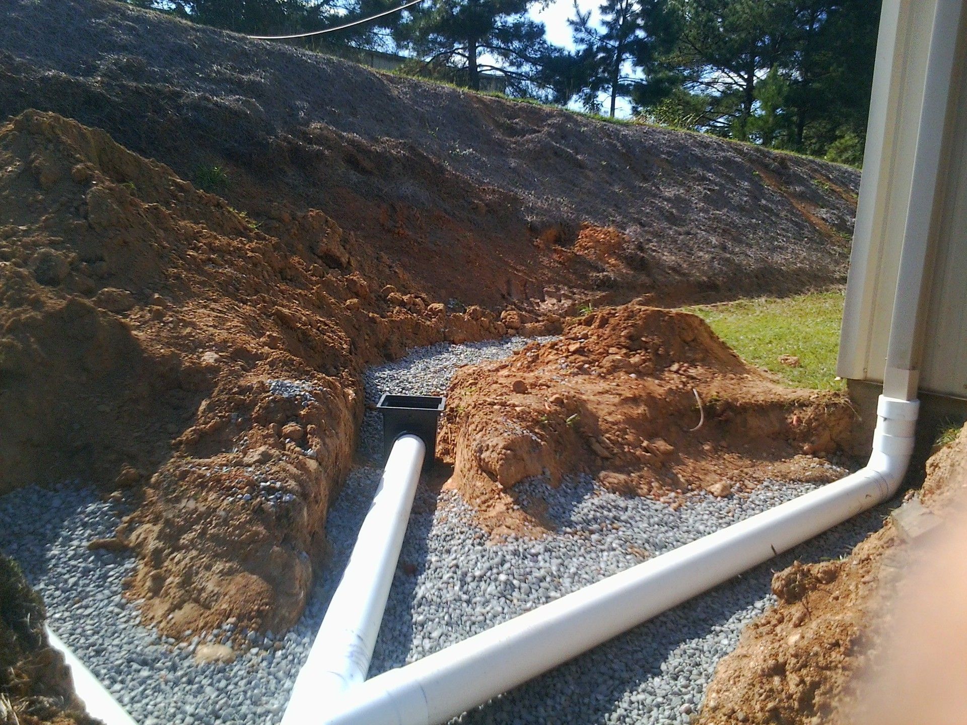 Construction site with drainage pipes in a gravel bed, soil mounds, and hillside in the background.