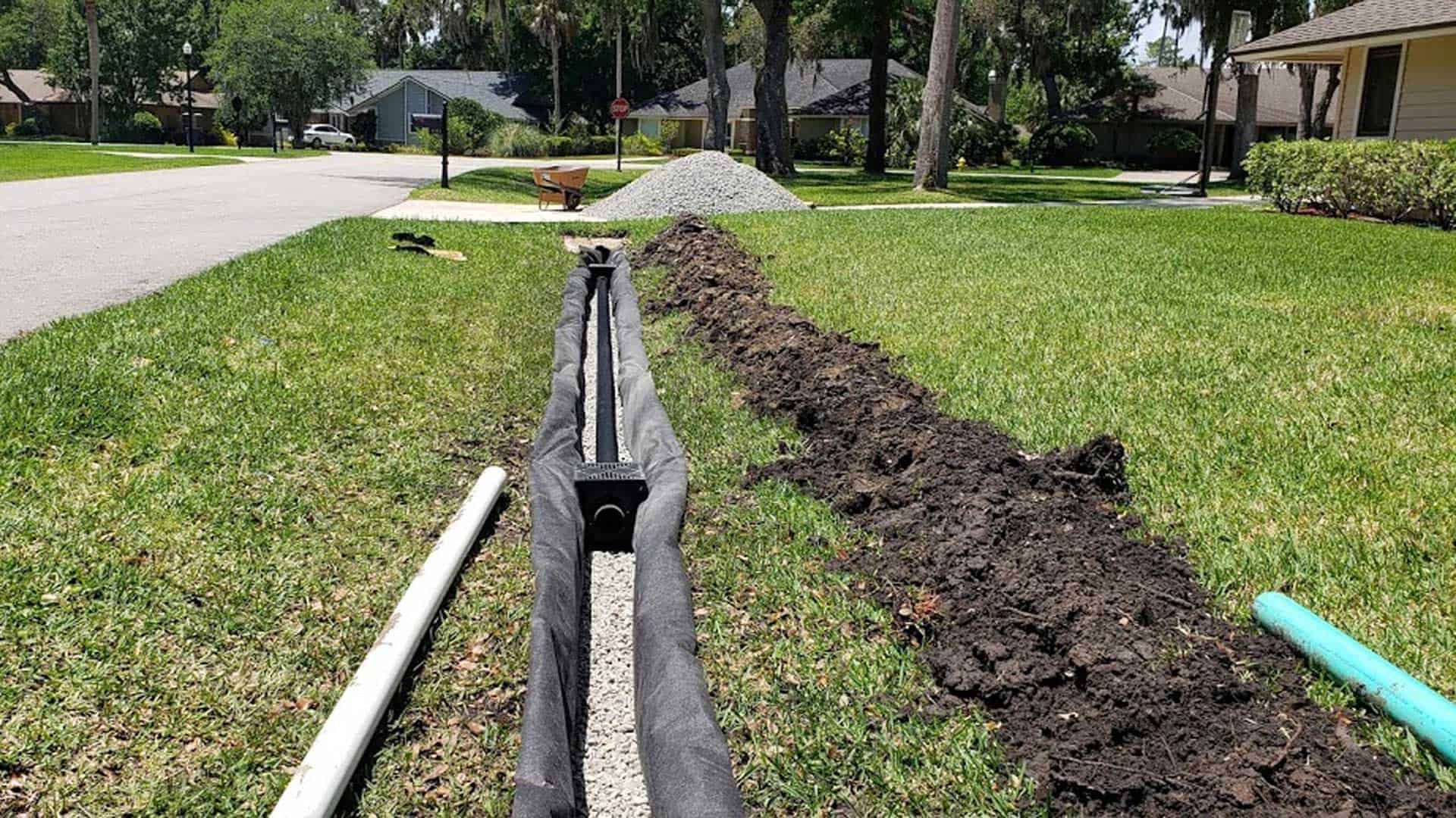 A trench drain being installed in a grassy yard, with gravel and pipes visible.