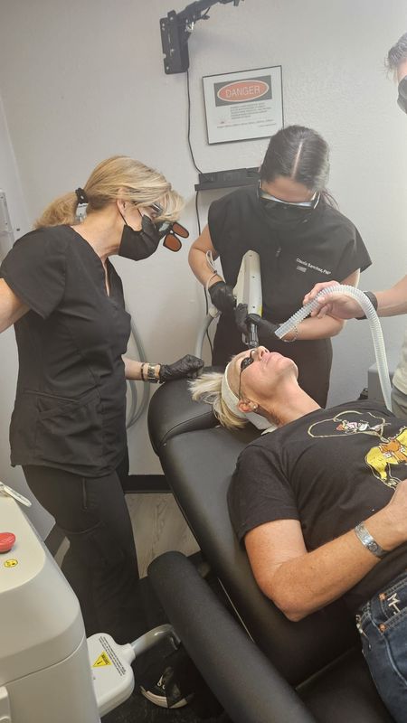 Three people in a medical setting. Two staff in scrubs, one treating a patient lying down.