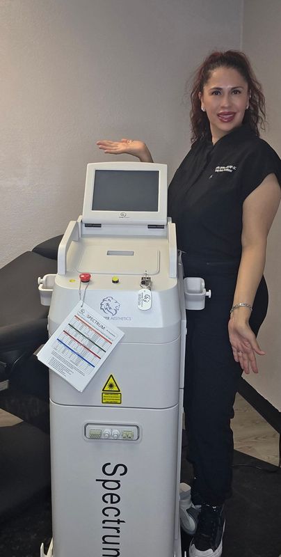 Woman in black next to a white Spectrum laser machine, smiling, in a medical office.