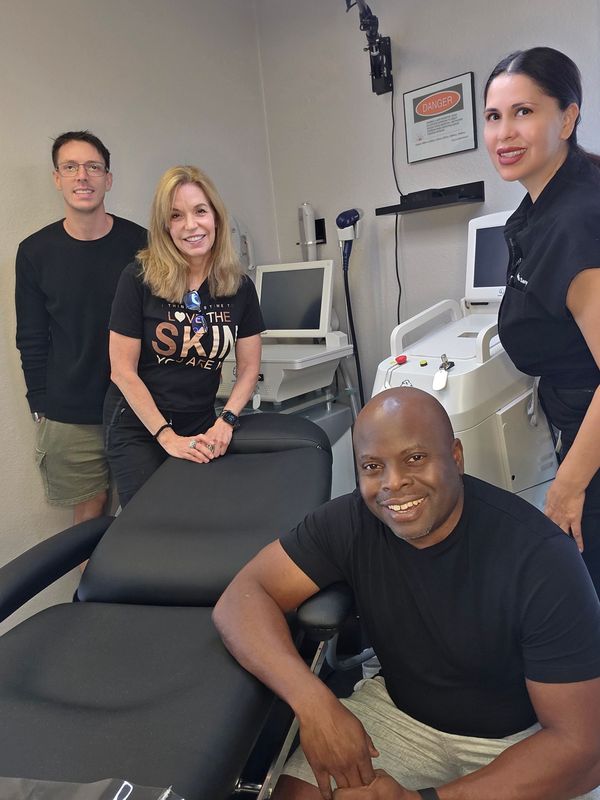 Four people in a clinic setting; a man sits on a medical bed smiling, others stand and smile near equipment.