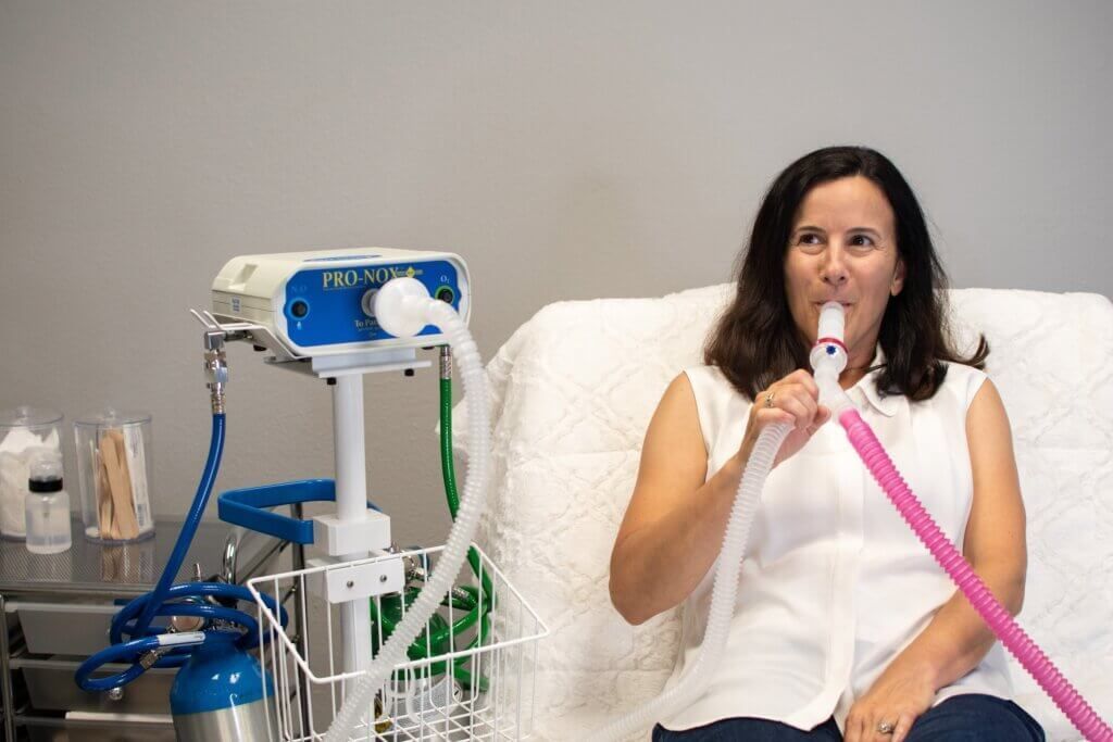 Woman using a respiratory device indoors, seated on a bed. Pink and white tube, blue machine.