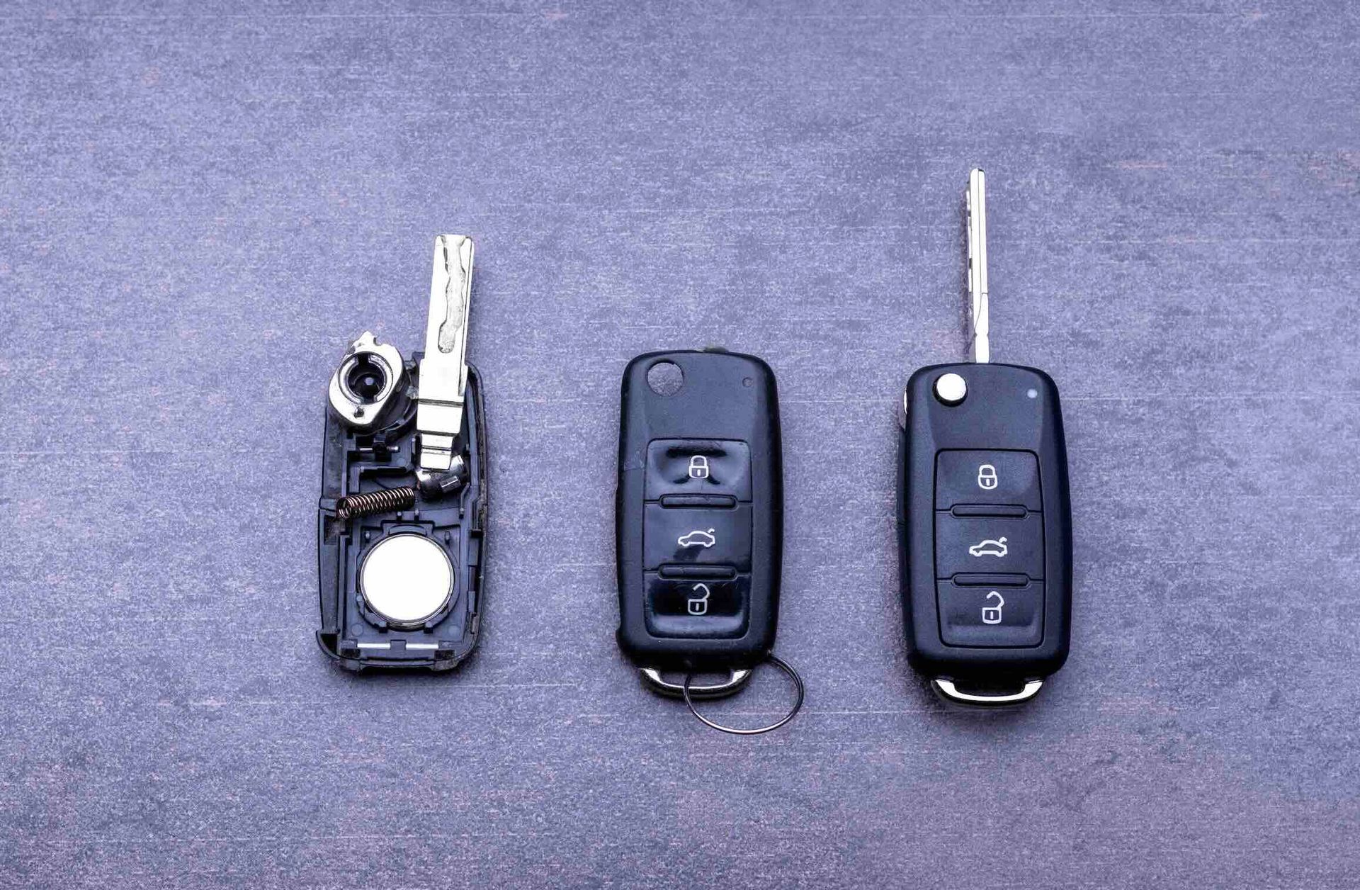 Three black car key fobs on a gray surface: one disassembled showing internal parts, and two assembled with metal keys.