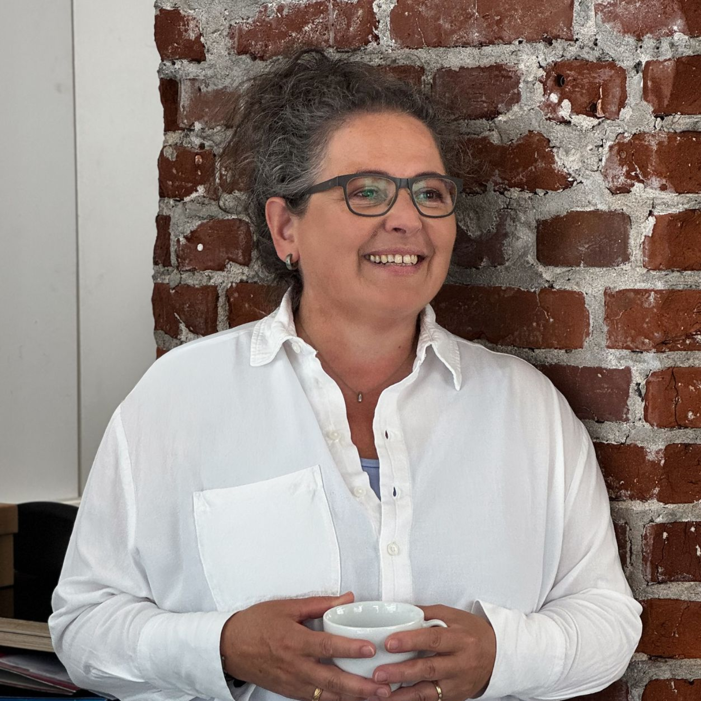 Frau mit Brille, lächelnd, Becher haltend, an Ziegelwand gelehnt.