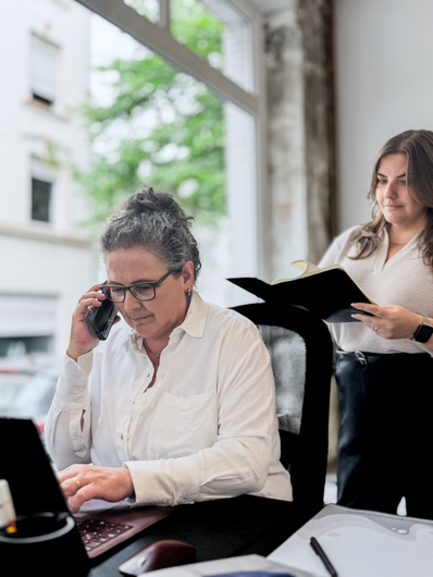 Frau telefoniert im Büro am Laptop, eine andere Frau steht in der Nähe und hält eine Akte.