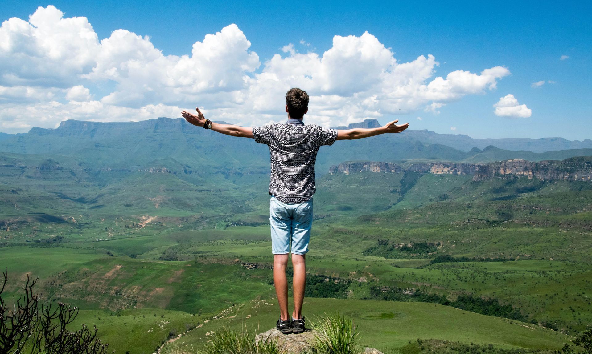 Mann mit ausgestreckten Armen auf einem Berggipfel mit Blick auf ein grünes Tal und Berge unter einem blauen Himmel mit Wolken.