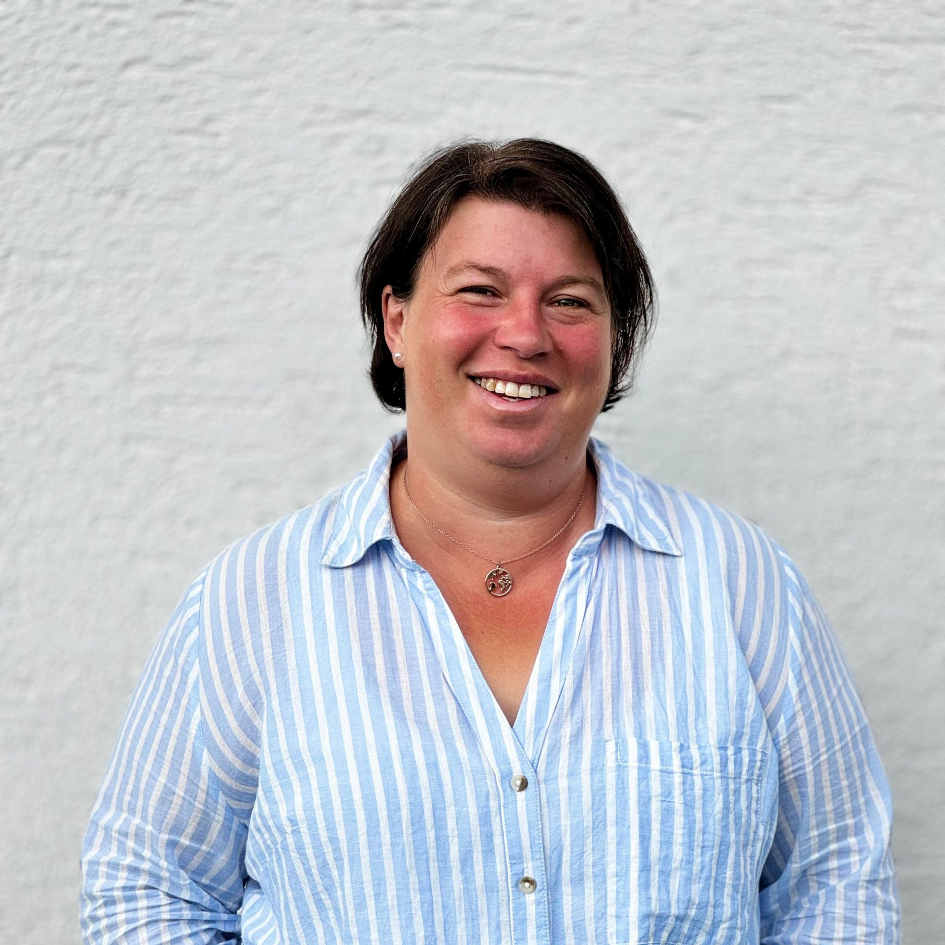 Frau mit dunklem Haar, lächelnd, trägt ein blau gestreiftes Hemd, steht vor einer weißen Wand.