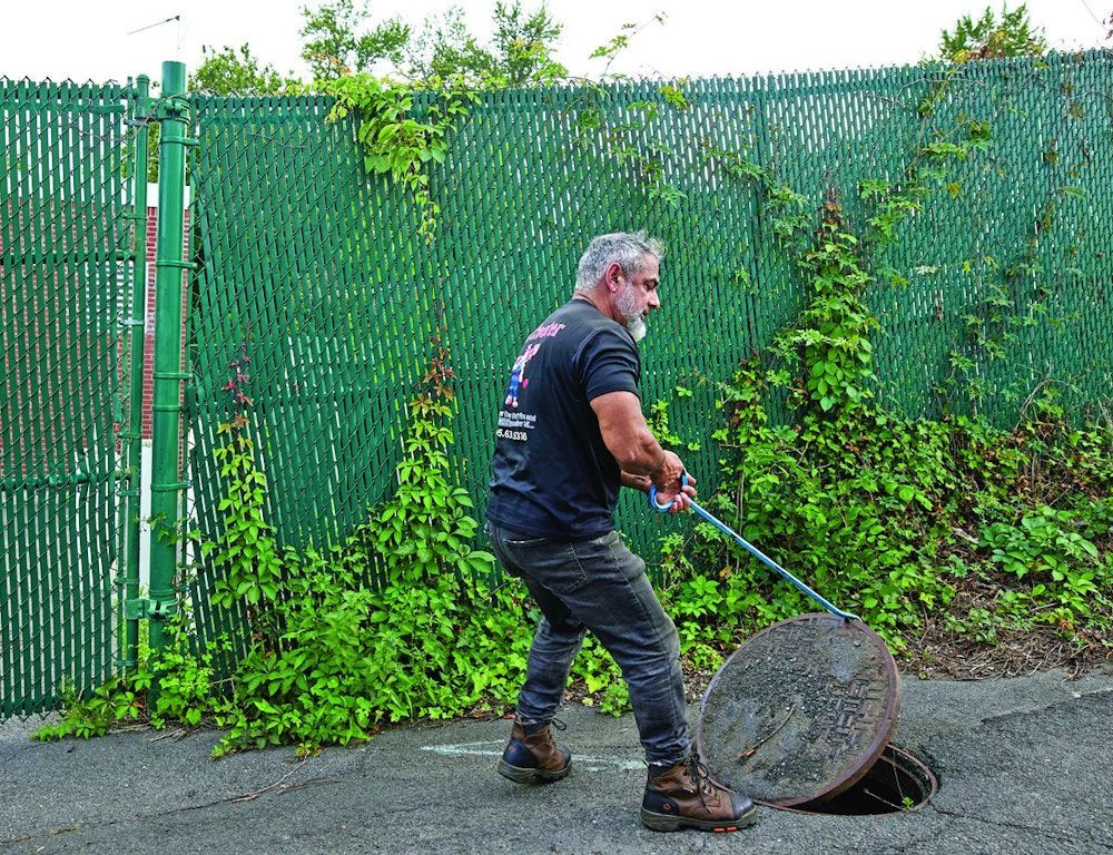 Man lifting a manhole cover on a street, beside a green fence covered in vines.